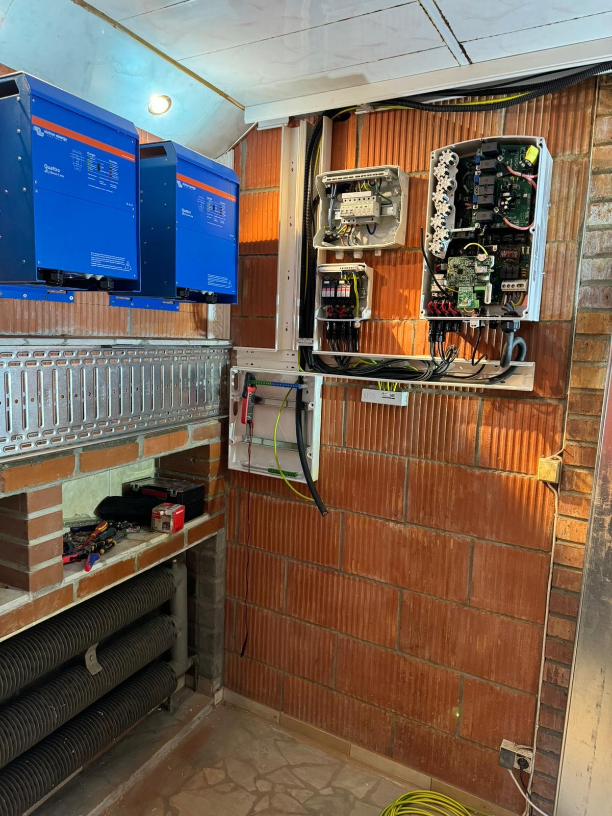 Instalacja paneli fotowoltaicznych w pomieszczeniu z ceglaną ścianą, widoczne inwertery w niebieskich obudowach, otwarte puszki elektryczne z okablowaniem i narzędzia na półce