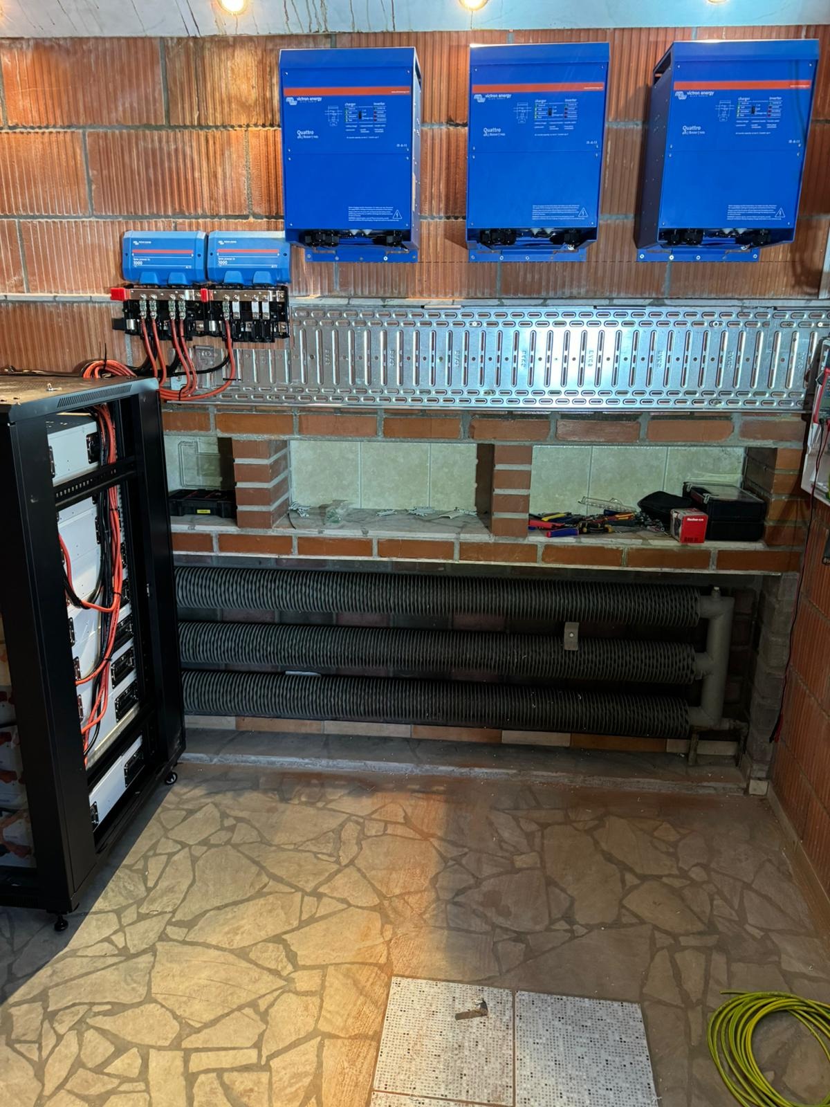 Instalacja fotowoltaiczna w pomieszczeniu: niebieskie inwertery na ceglanej ścianie, szafa rack z bateriami, kable, peszle i narzędzia na półce.