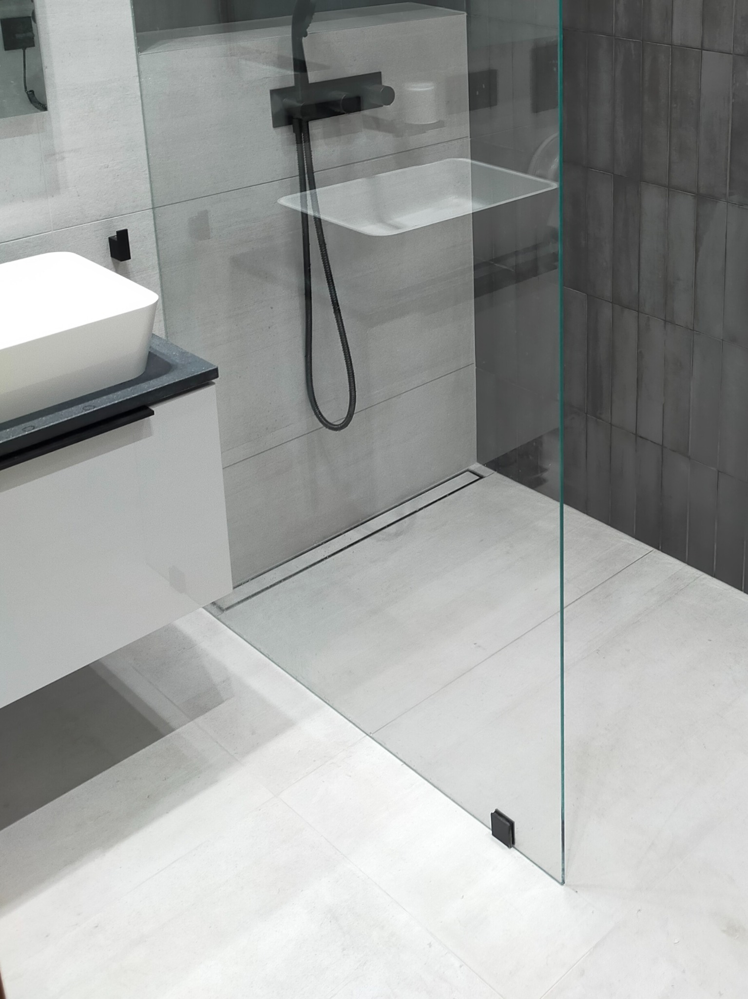 Nowoczesna łazienka z kabiną prysznicową typu walk-in, szare płytki na ścianie i podłodze, czarna armatura, biała umywalka nablatowa na szafce.