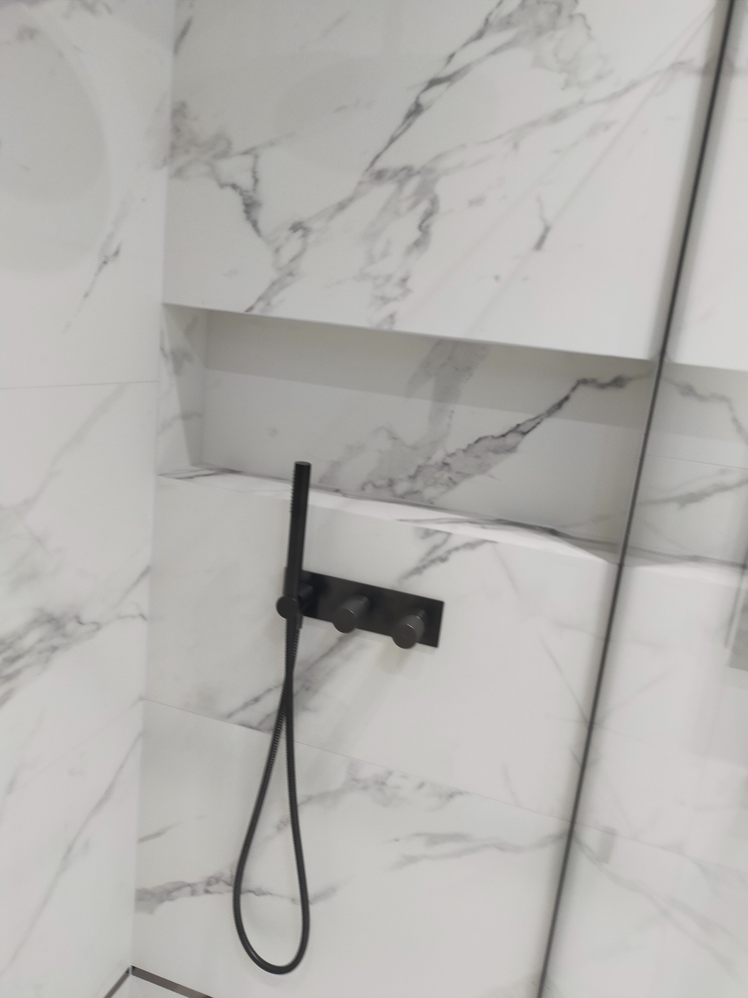 Ściana kabiny prysznicowej wyłożona białymi płytkami imitującymi marmur, z wnęką i czarną armaturą.