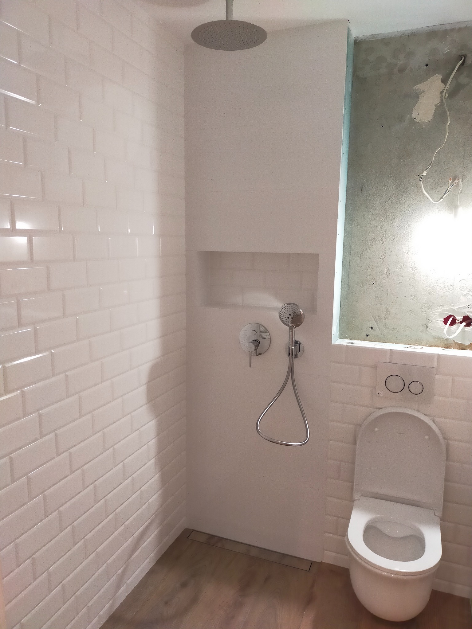 Nowoczesna łazienka w trakcie remontu: białe, błyszczące płytki typu metro na ścianach, deszczownica, armatura chromowana, toaleta z otwartą klapą i surowa ściana z widocznymi przewodami.