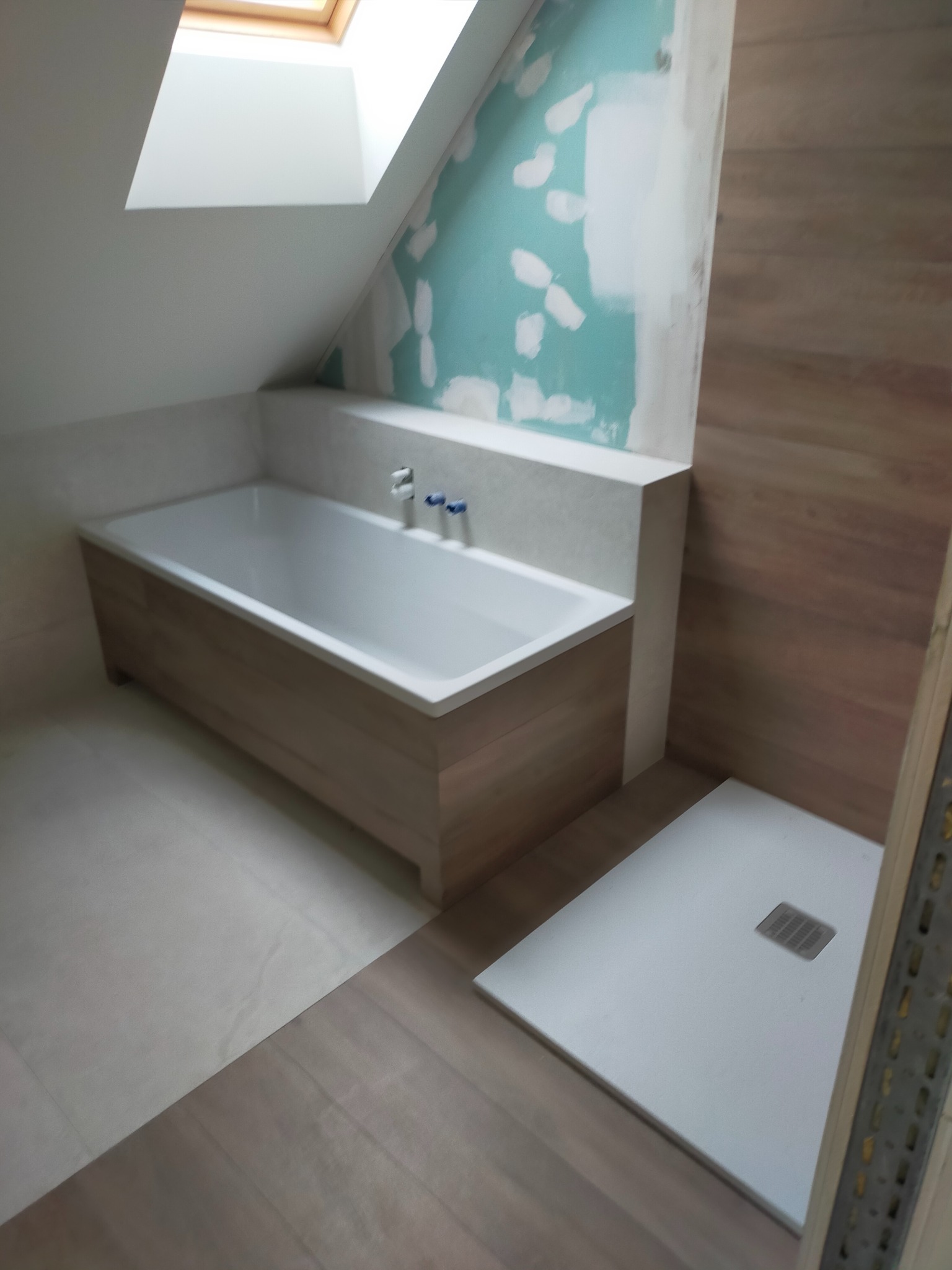 Wnętrze łazienki na poddaszu w trakcie prac wykończeniowych: zabudowana wanna z drewnopodobnym wykończeniem, biały brodzik z odpływem liniowym, fragment ściany z surowym tynkiem i fragment...