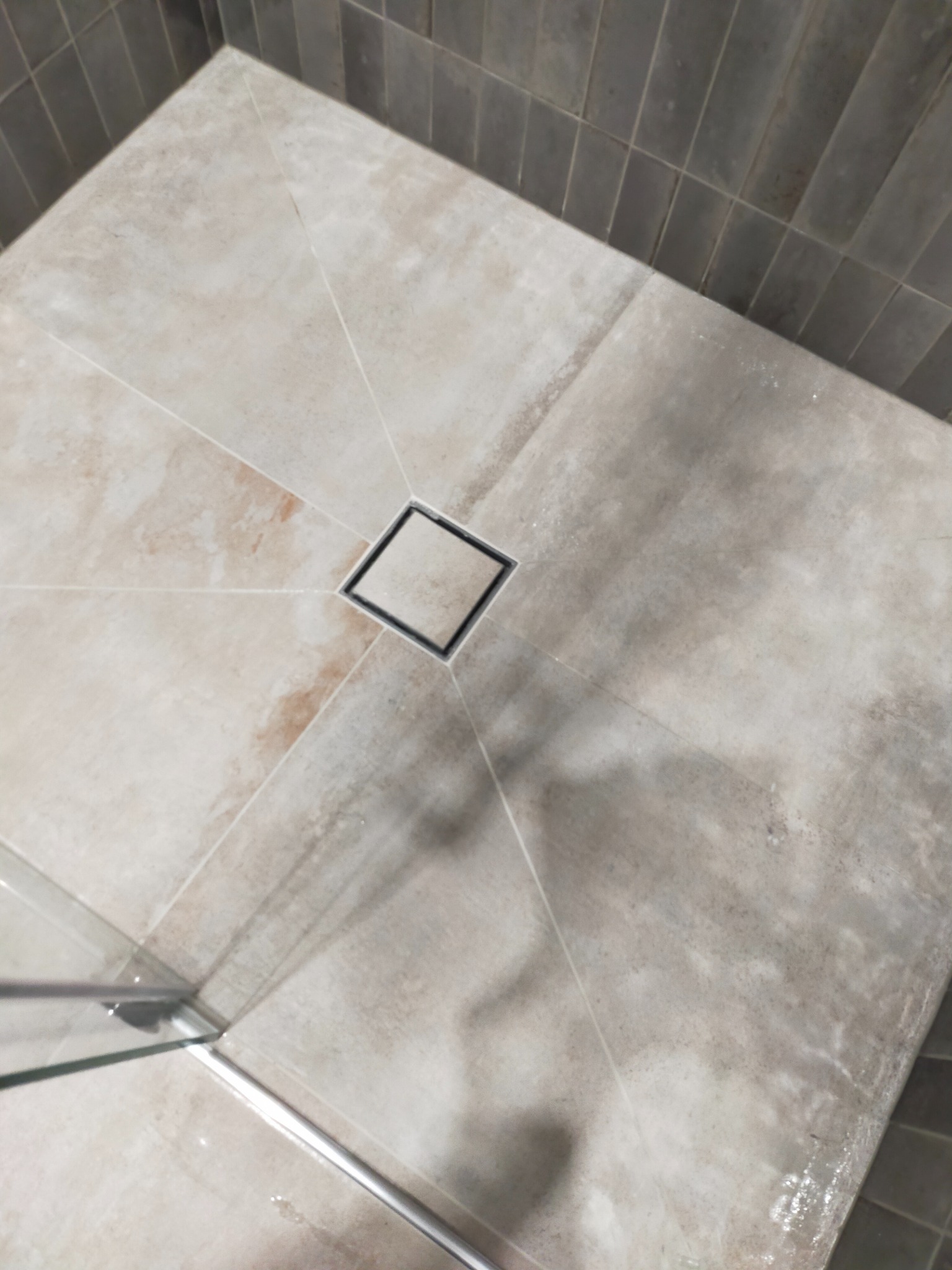 Beżowe płytki podłogowe w kabinie prysznicowej z centralnie umieszczonym odpływem liniowym w kształcie kwadratu, ciemnoszare płytki na ścianie w tle, widoczny fragment szklanej ścianki kabiny.