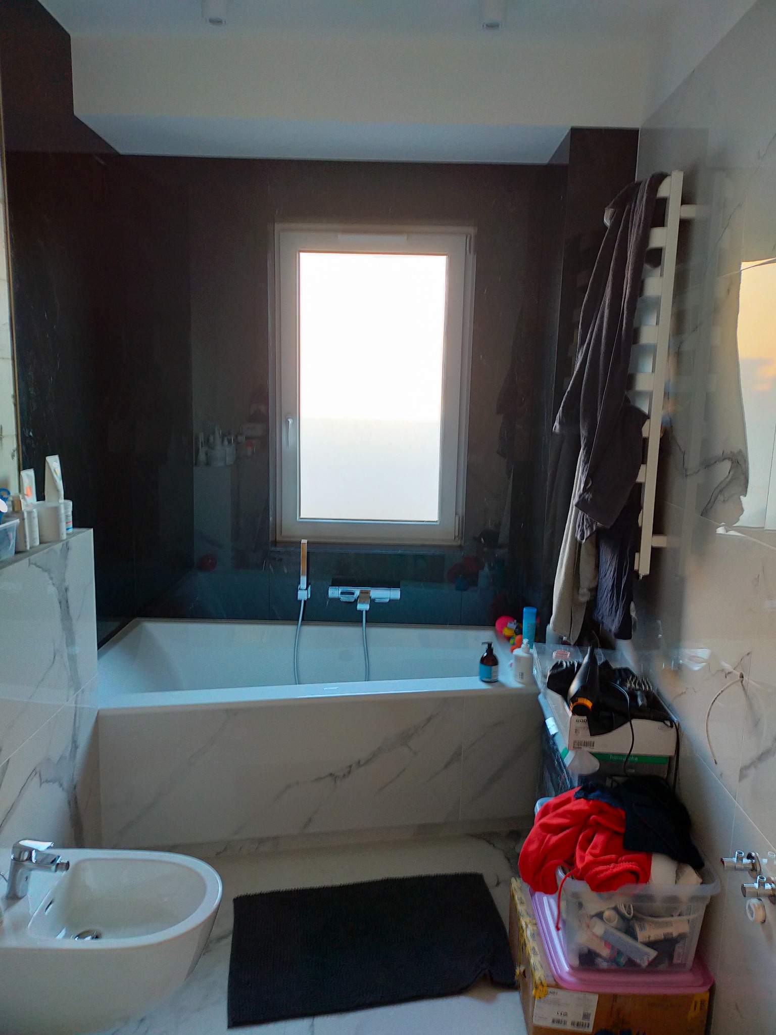 Wnętrze łazienki z wanną obudowaną marmurem, bidetem i grzejnikiem drabinkowym, na tle ciemnych płytek, z widocznymi kosmetykami i akcesoriami w nieładzie.