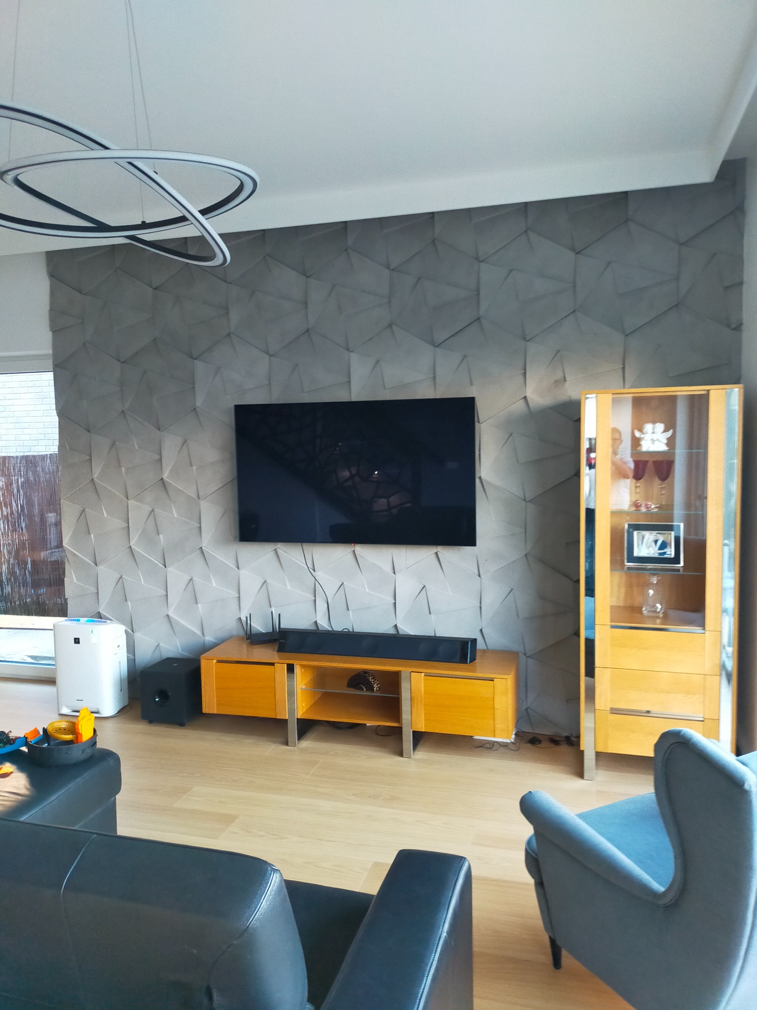 Salon z szarą ścianą z geometrycznym wzorem 3D, telewizorem, szafką RTV z głośnikiem soundbar, witryną, nowoczesną lampą wiszącą i fragmentami mebli wypoczynkowych.