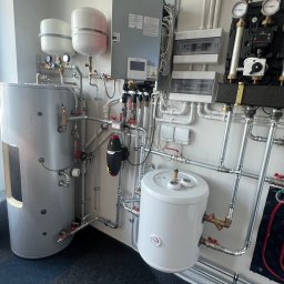 FT GLOBAL SPÓŁKA Z OGRANICZONĄ ODPOWIEDZIALNOŚCIĄ - Zestaw urządzeń w kotłowni: szary zbiornik akumulacyjny z wizjerem, biały zasobnik CWU, rozdzielacz grzewczy z manometrami, skrzynka elektryczna z ostrzeżeniem o napięciu, orurowanie ze złączkami.