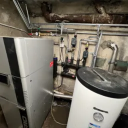 Srebrna pompa ciepła Evopell z podłączonym zasobnikiem Kospel SW-100 w kotłowni z rurami instalacji grzewczej na ścianie.