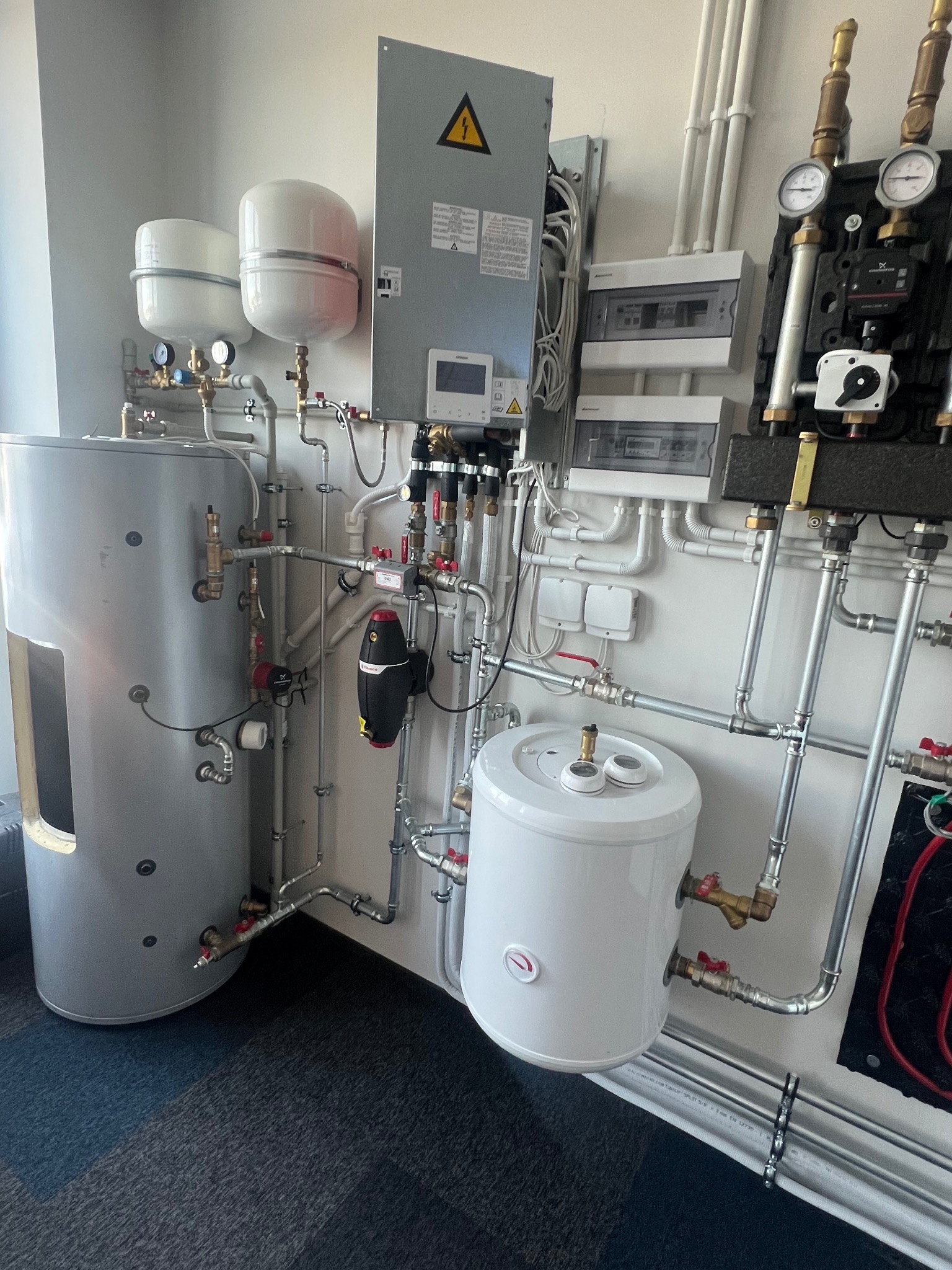 Zestaw urządzeń w kotłowni: szary zbiornik akumulacyjny z wizjerem, biały zasobnik CWU, rozdzielacz grzewczy z manometrami, skrzynka elektryczna z ostrzeżeniem o napięciu, orurowanie ze złączkami.