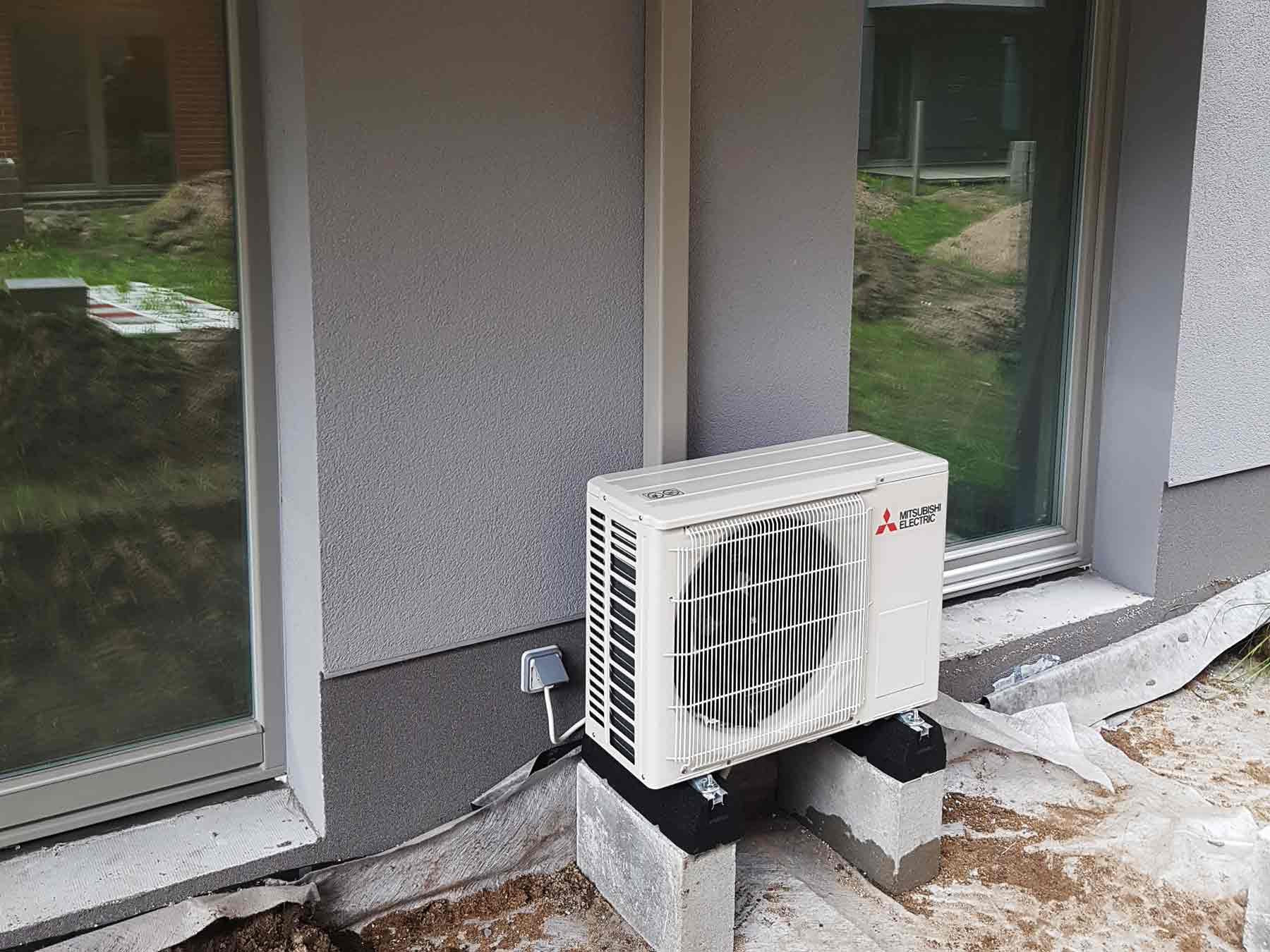 Zewnętrzna jednostka klimatyzacji Mitsubishi Electric, umieszczona na betonowych bloczkach przy ścianie budynku z widocznym oknem i fragmentem trawnika.