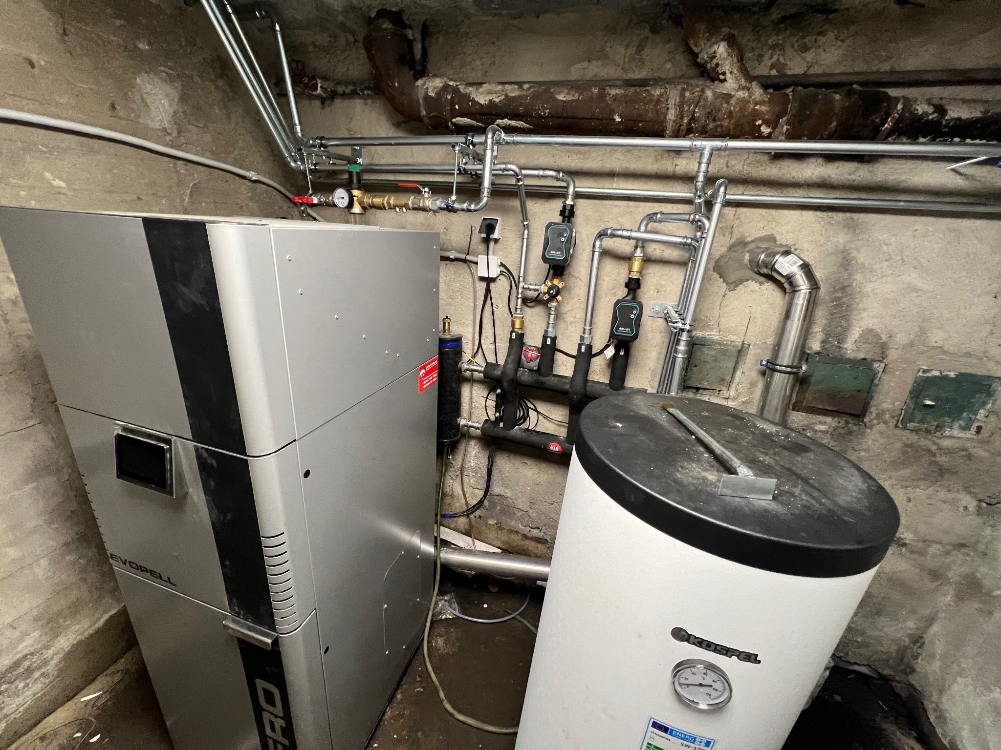Srebrna pompa ciepła Evopell z podłączonym zasobnikiem Kospel SW-100 w kotłowni z rurami instalacji grzewczej na ścianie.