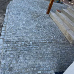 Ułożona kostka granitowa tworząca ścieżkę z łukowatym wzorem, przylegająca do betonowych schodów i drewnianego słupa. Widoczny trawnik w tle.