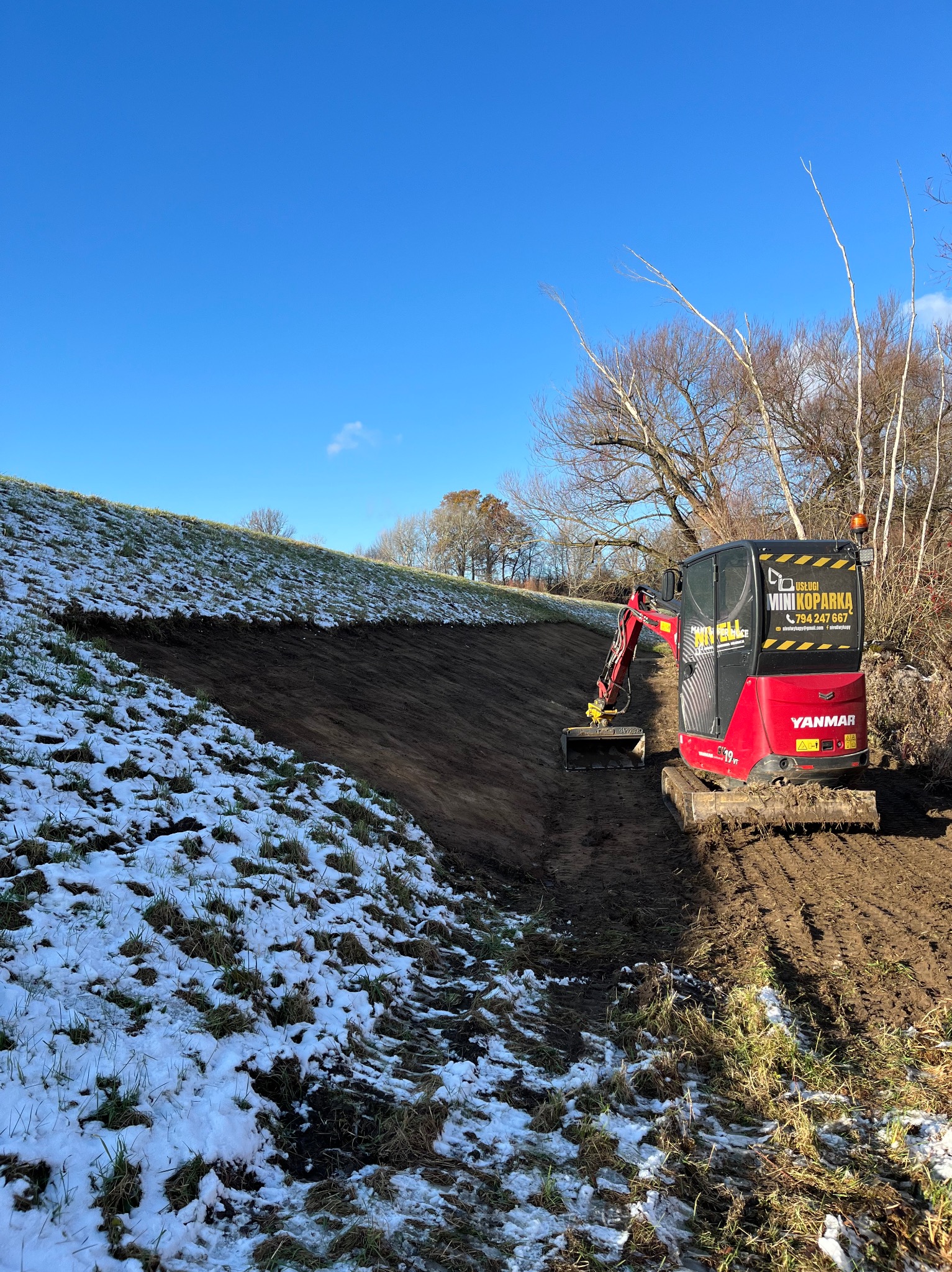 Czerwona mini koparka Yanmar na zboczu z resztkami śniegu, wykonująca wykop w ziemi, widoczne ślady gąsienic i łyżki, błękitne niebo w tle.