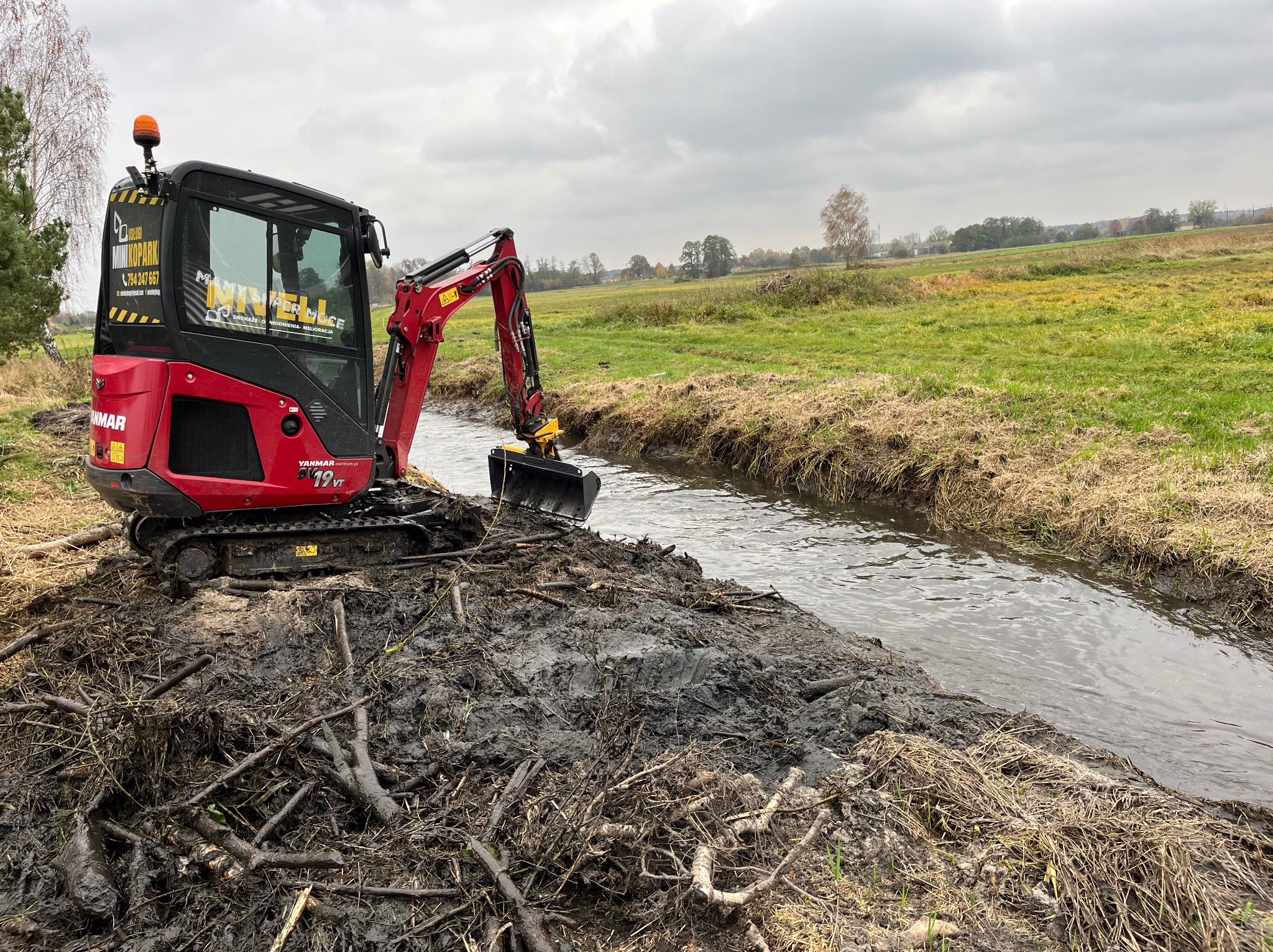 Czerwona mini koparka Yanmar 19VT na brzegu rowu melioracyjnego, usuwająca gałęzie i muł, z widocznym logo firmy 'Mini Koparki' na kabinie, w tle zielone pola i pochmurne niebo.