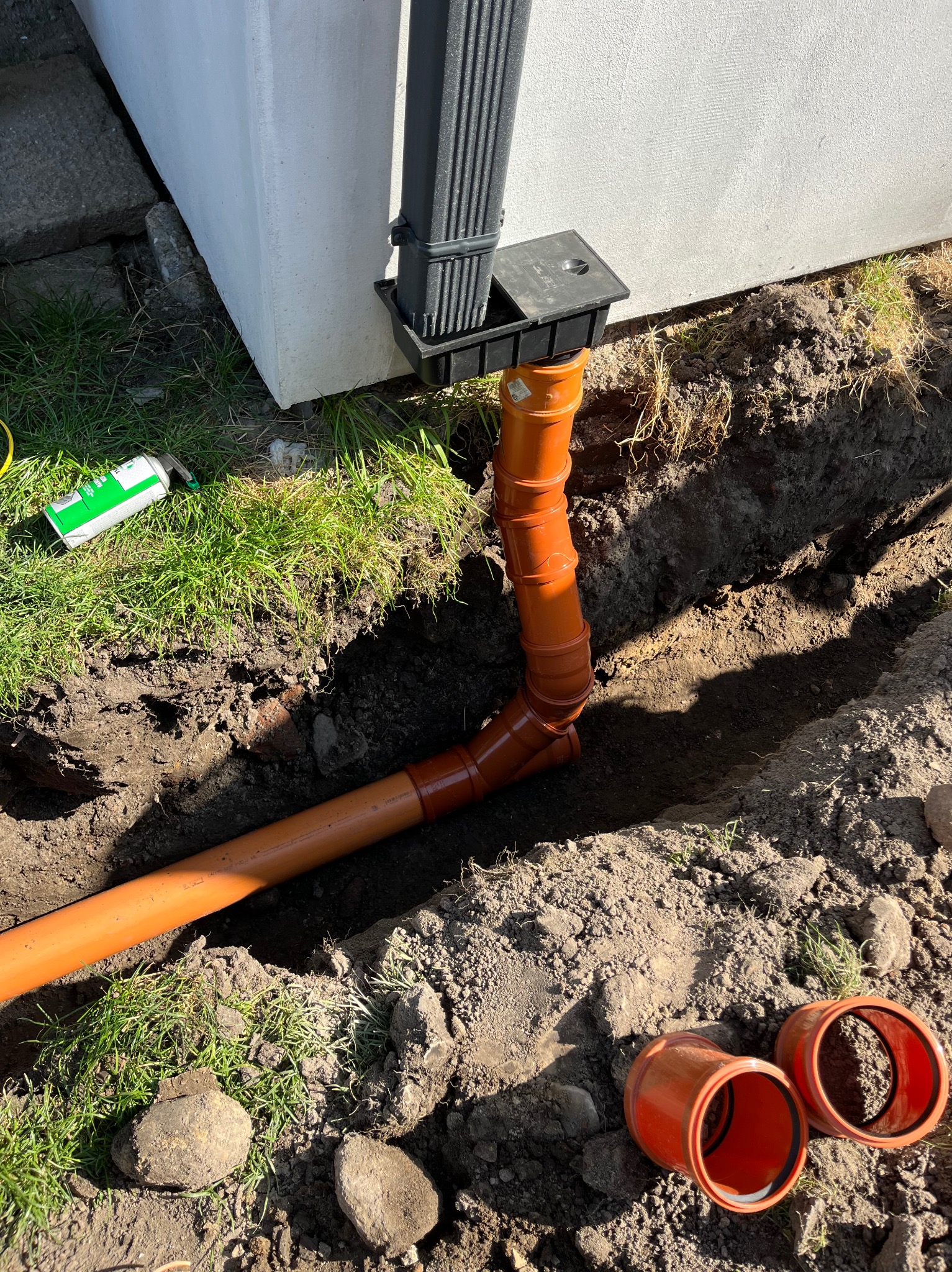 Pomarańczowe rury kanalizacyjne ułożone w wykopie ziemnym, połączone z czarną rynną spustową przy fundamencie budynku, z widocznymi złączkami i narzędziem na trawie.