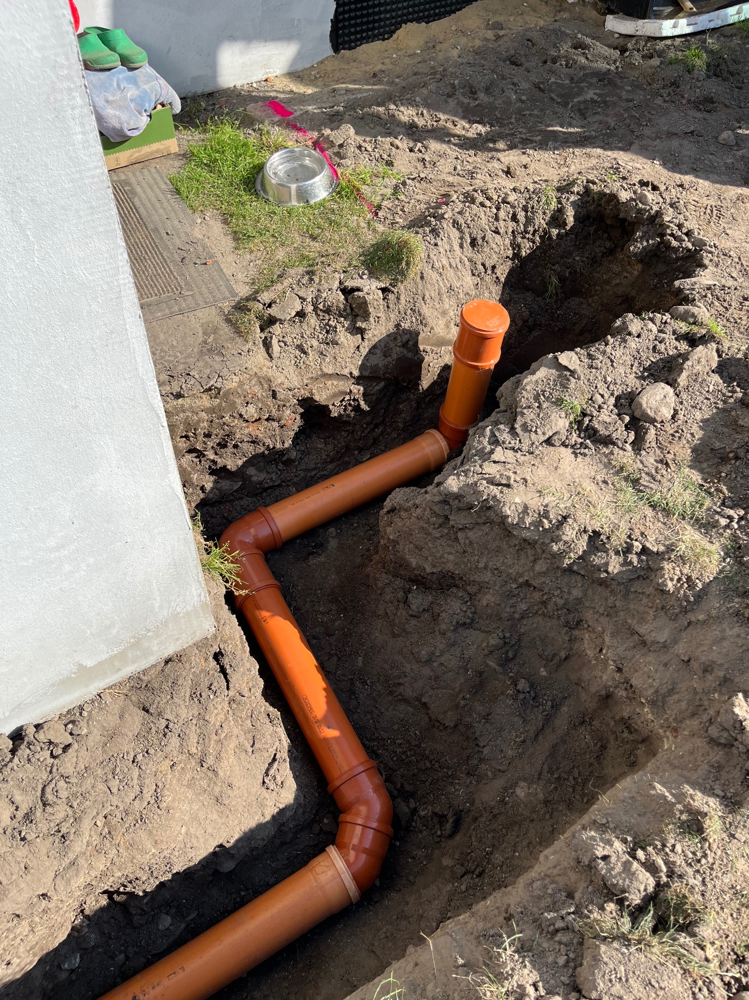 Pomarańczowe rury kanalizacyjne ułożone w wykopie obok fundamentu budynku, widoczne złącza i pionowy odcinek, obok leżą zielone kalosze i miska.