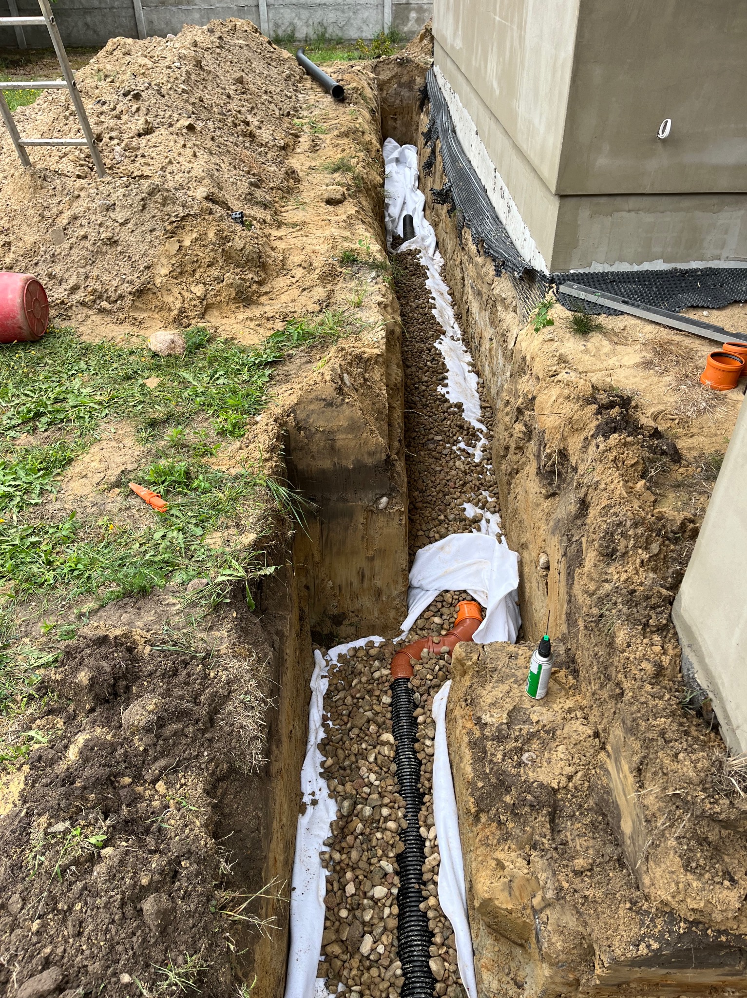 Wykop z ułożoną rurą drenarską w geowłókninie, obsypany kamieniami, widoczne elementy instalacji przy fundamencie budynku.