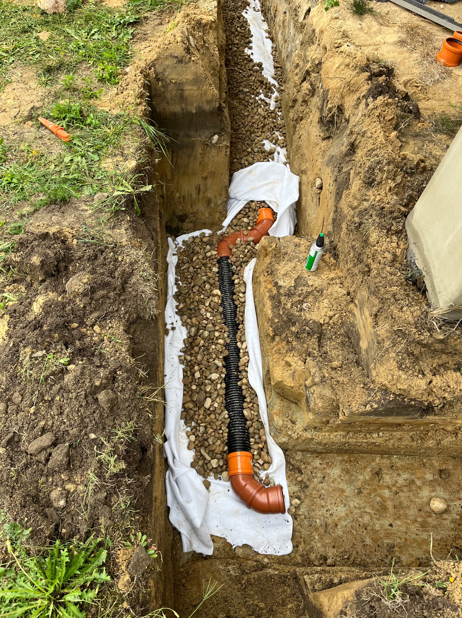 Wykop pod drenaż z rurami drenarskimi w geowłókninie obsypany drobnym żwirem, widoczny fragment fundamentu i narzędzie w tle.