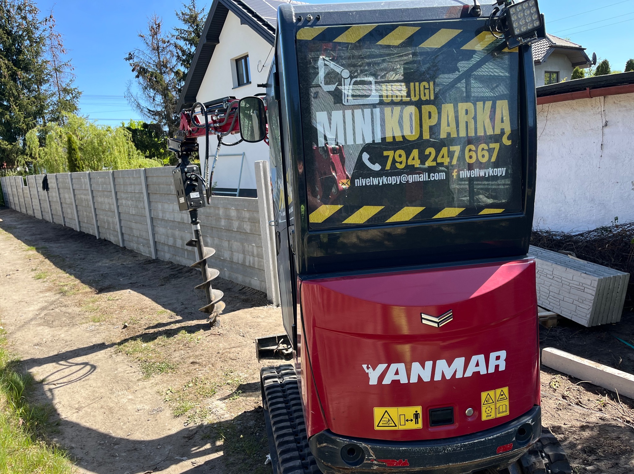Czerwona mini koparka Yanmar z wiertłem obok betonowego ogrodzenia panelowego, w tle dom jednorodzinny i błękitne niebo. Widoczne logo firmy na maszynie.