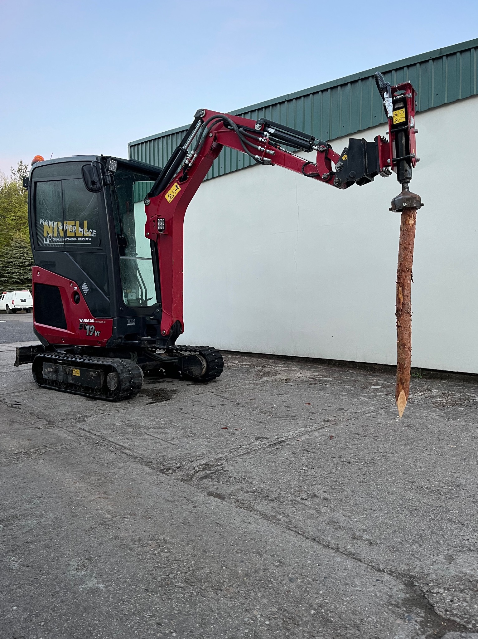 Czerwony minikoparka Yanmar wbija pal drewniany w ziemię. Widoczny budynek w tle. Maszyna gotowa do pracy w terenie.