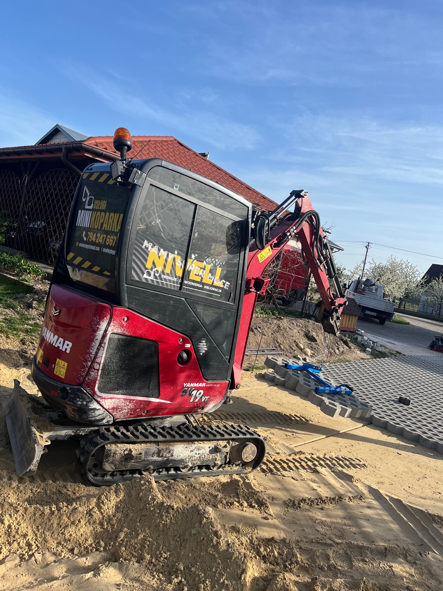 Czerwona mini koparka Yanmar na piasku obok ułożonej kostki brukowej. Na kabinie naklejka z napisem NIVELLE i numerem telefonu. Widok z boku, w tle dom i błękitne niebo.