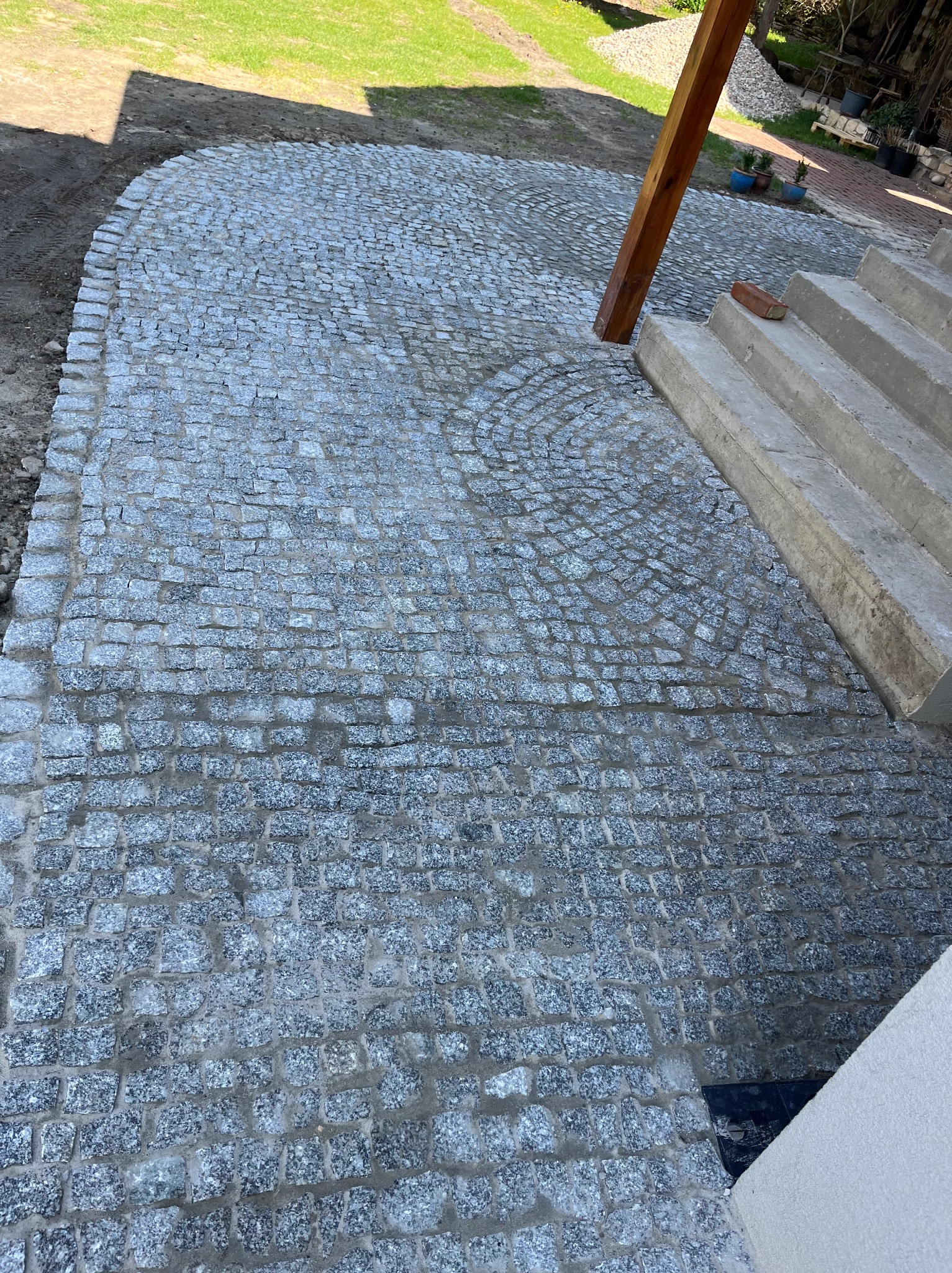 Ułożona kostka granitowa tworząca ścieżkę z łukowatym wzorem, przylegająca do betonowych schodów i drewnianego słupa. Widoczny trawnik w tle.