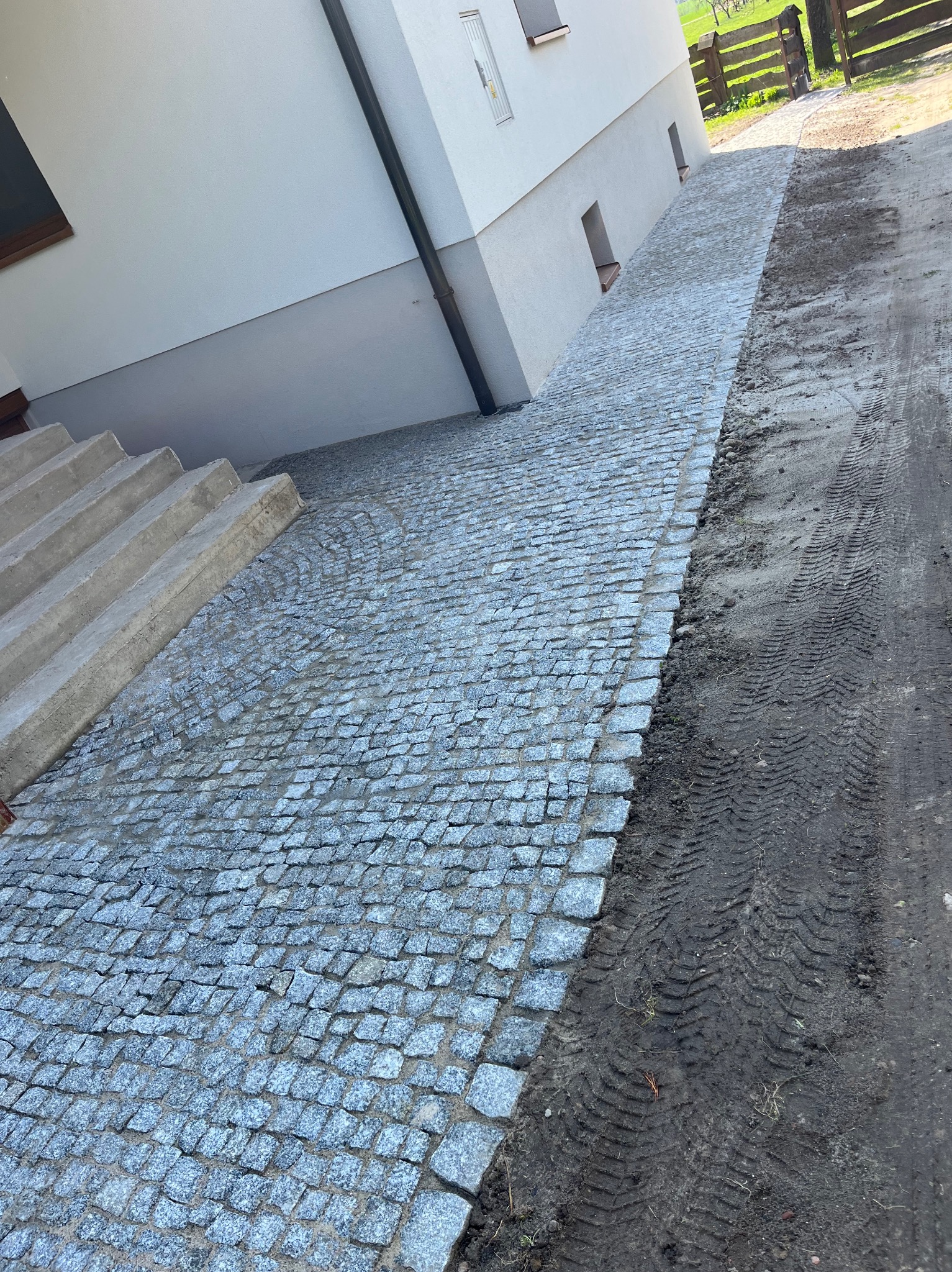 Granitowa kostka brukowa ułożona w łukowaty wzór przed budynkiem, z widocznymi śladami opon na ziemi obok, sugerująca etap prac wykończeniowych.