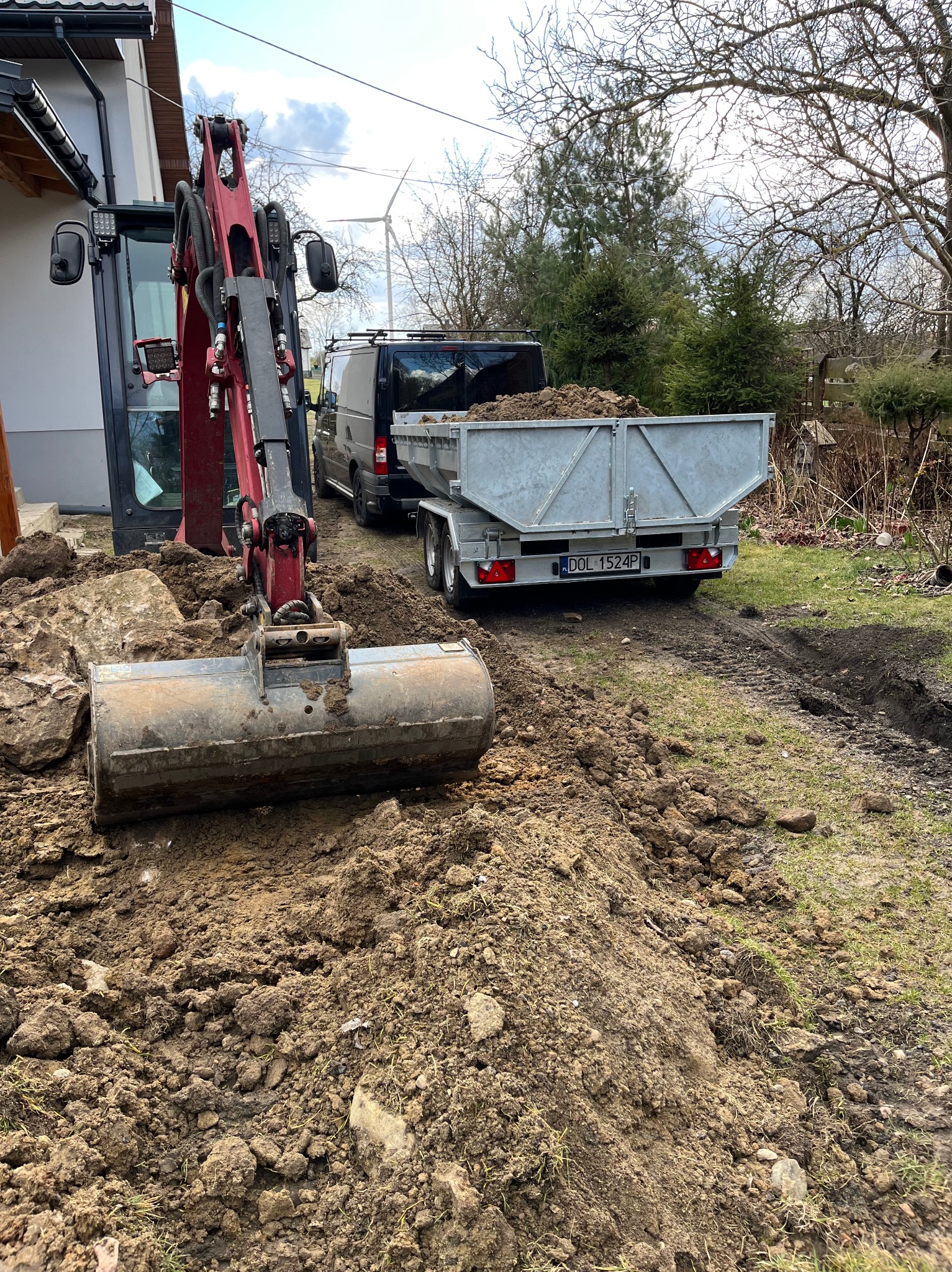 Czerwony minikoparka w trakcie prac ziemnych, z przyczepą pełną ziemi w tle. Widoczny wykop i świeżo naruszona gleba. Na horyzoncie turbina wiatrowa.