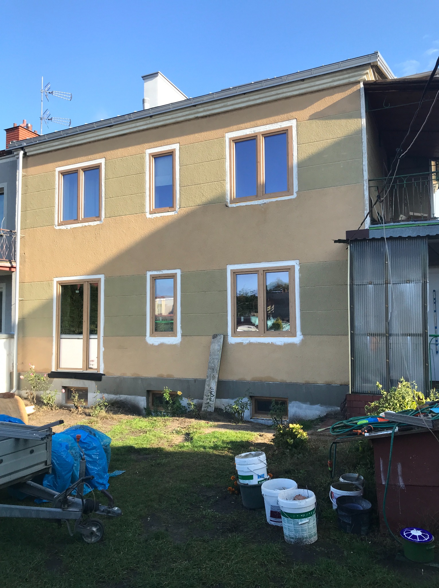 Dwupiętrowy dom z nowymi oknami z drewnianymi ramami, widoczne ślady po montażu okien, teren przed domem z elementami budowlanymi i sprzętem ogrodniczym.