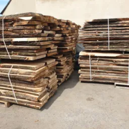 Dwa stosy świeżo przetartej tarcicy z widoczną korą na krawędziach, ułożone na paletach i zabezpieczone sznurkiem, na betonowym podłożu, w tle budynek.