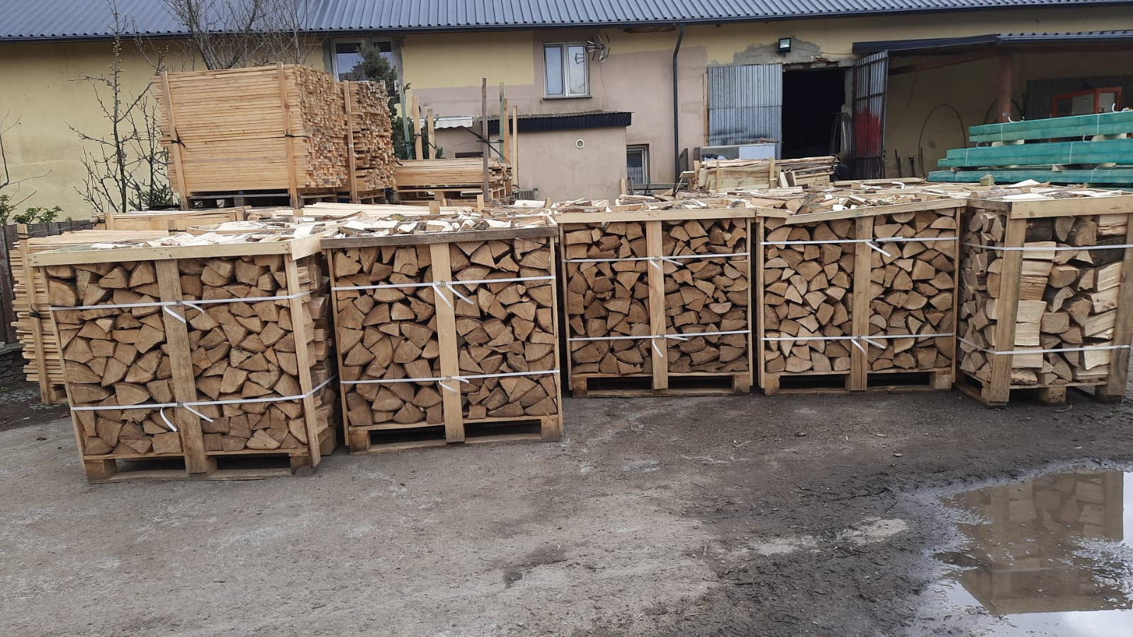 Ułożone w drewnianych skrzyniach na paletach pocięte i porąbane kawałki drewna kominkowego, zabezpieczone białym sznurkiem, na tle budynków gospodarczych i stosu tarcicy.