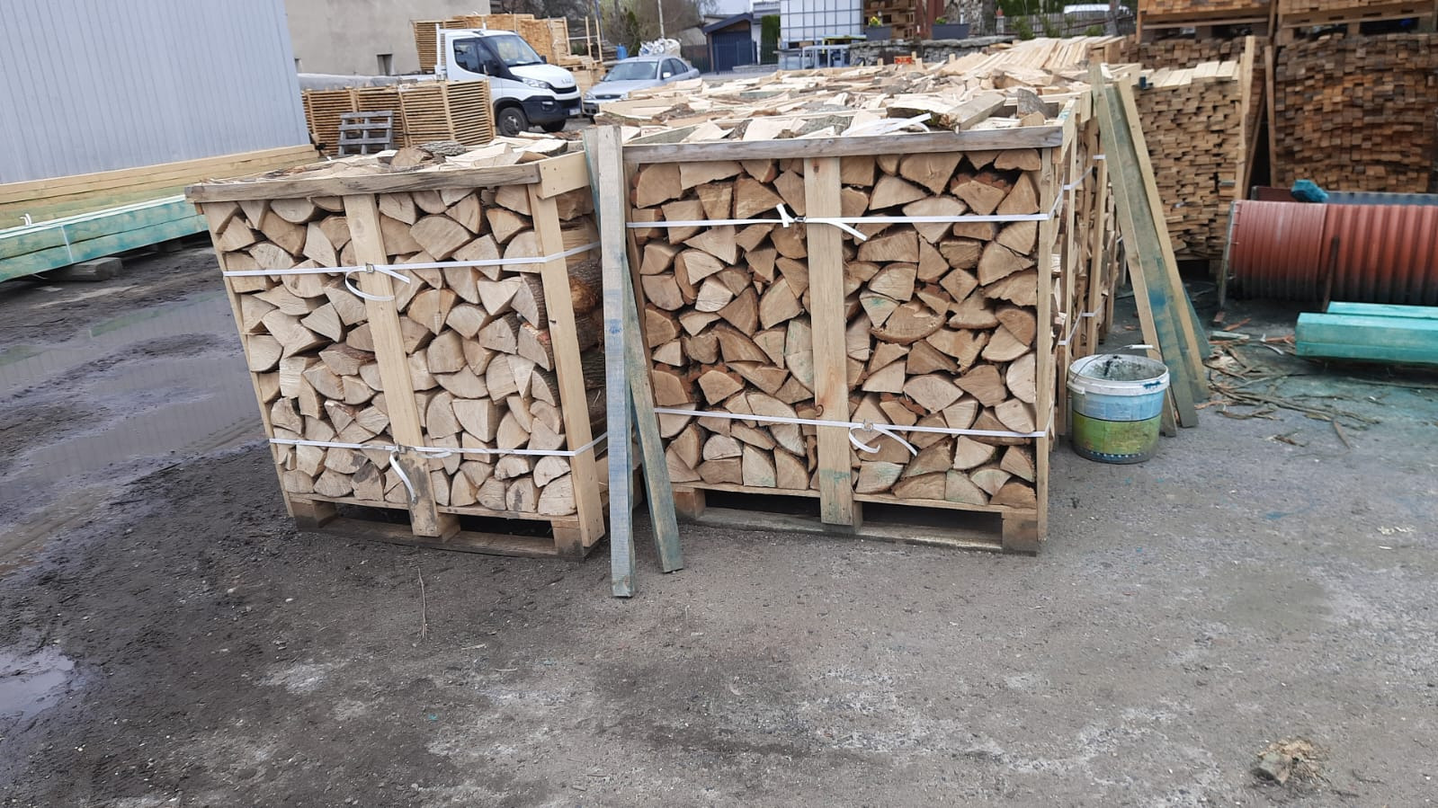 Dwa kontenery paletowe wypełnione pociętym drewnem kominkowym, zabezpieczone białymi taśmami, na zewnątrz, obok widoczne deski i metalowe beczki.