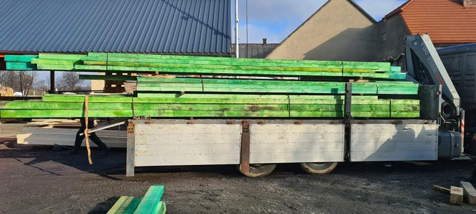 Stos impregnowanej na zielono tarcicy budowlanej, przewożony na platformie samochodu ciężarowego z dźwigiem hydraulicznym, na tle zabudowań gospodarczych.