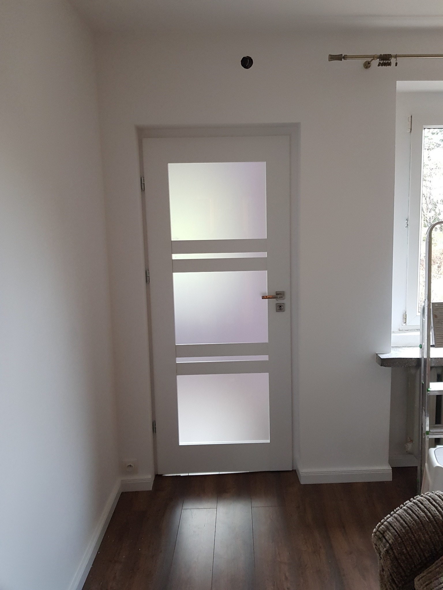 Białe, przeszklone drzwi wewnętrzne z trzema prostokątnymi szybami i złotą klamką, osadzone w świeżo pomalowanej na biało ścianie, widoczne na tle parkietu i fragmentu drabiny malarskiej przy oknie.