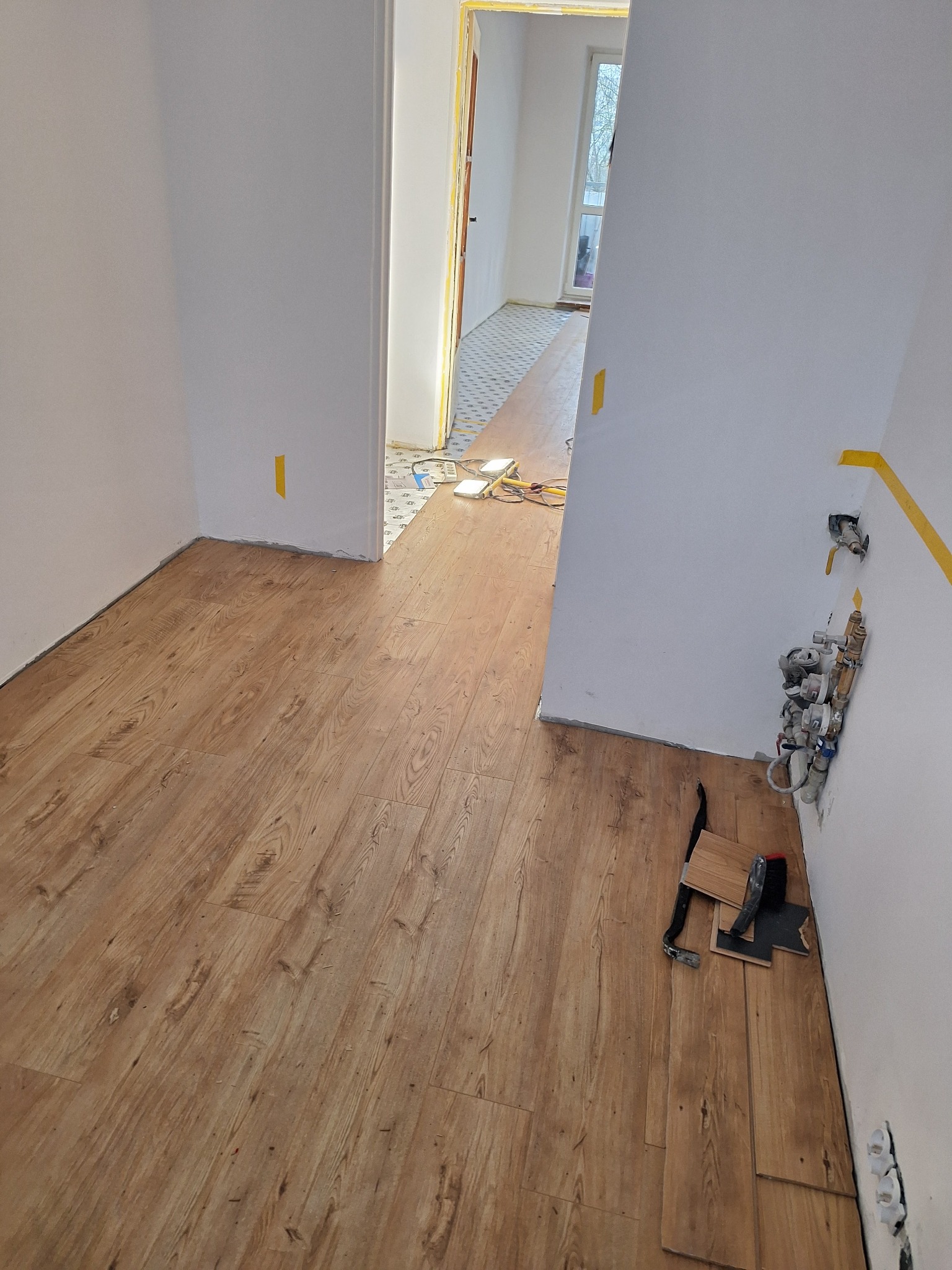 Układanie paneli podłogowych w jasnym pomieszczeniu z widocznymi narzędziami i wystającymi rurami wodnymi przy ścianie.