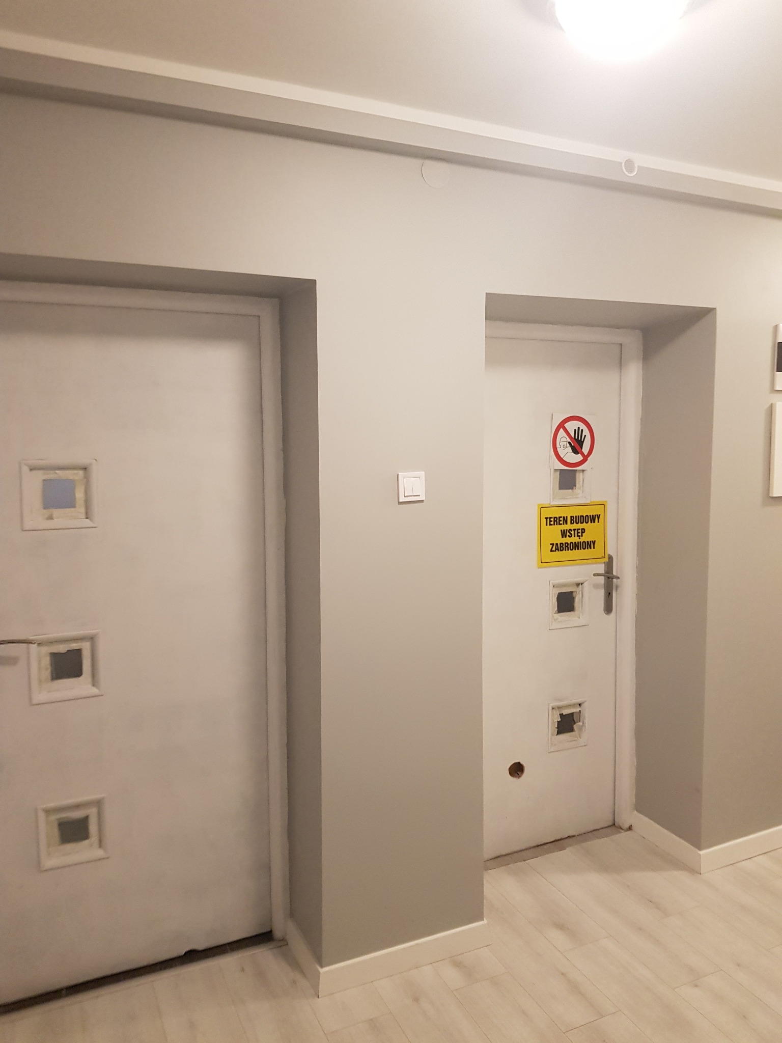 Dwa białe, uszkodzone drzwi w szarym korytarzu, z tabliczką ostrzegawczą 'Teren Budowy Wstęp Zabroniony' na jednych z nich, widoczne pozostałości po otworach inspekcyjnych.