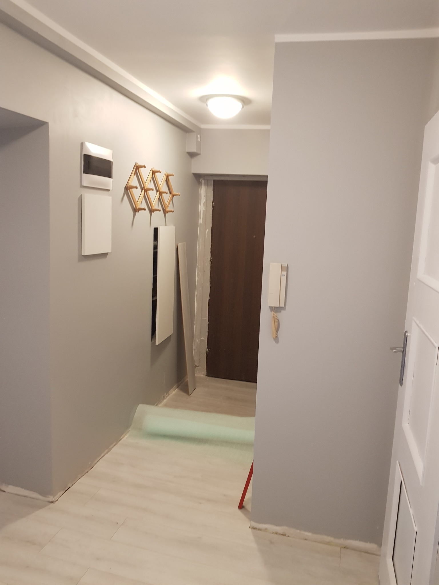 Świeżo pomalowany na szaro korytarz w mieszkaniu, widoczne akcesoria: wieszak na ubrania, domofon, skrzynka z bezpiecznikami i listwa przypodłogowa w trakcie montażu.