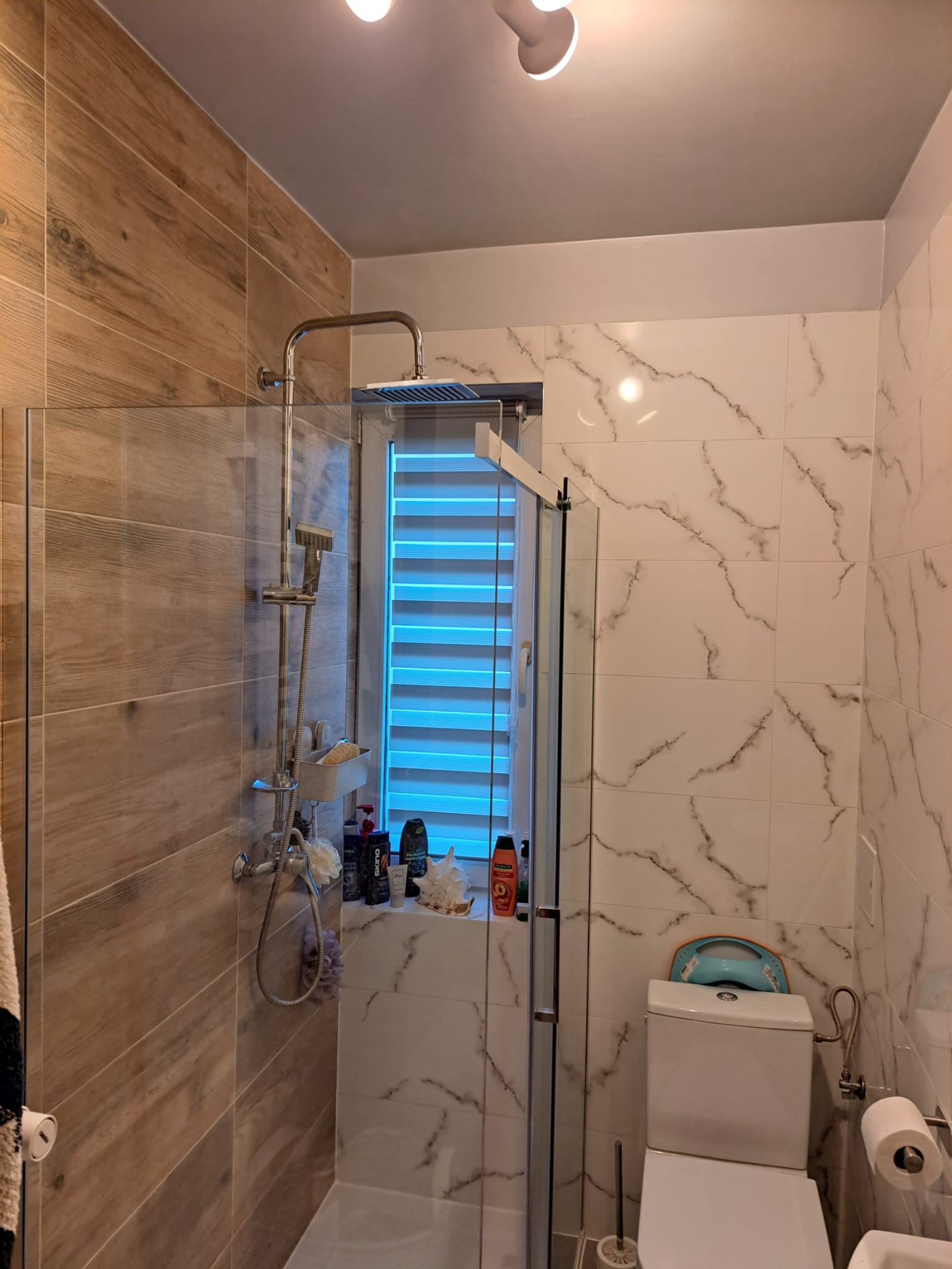 Nowoczesna łazienka z kabiną prysznicową typu walk-in, wykończona płytkami imitującymi drewno i marmur, z widoczną toaletą i oknem z żaluzjami poziomymi.