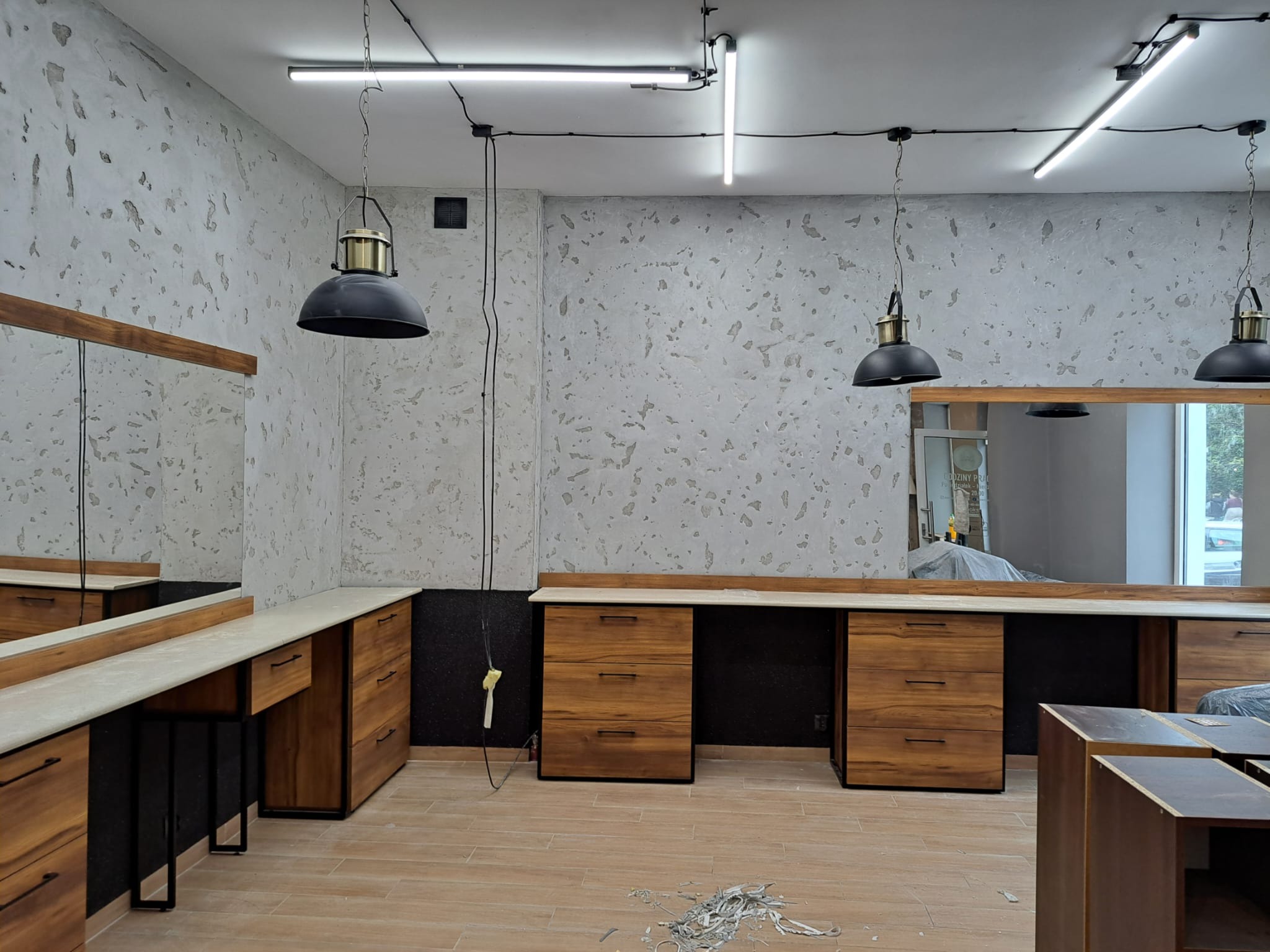 Wykończone wnętrze biurowe z drewnianymi szafkami, lustrami i industrialnymi lampami wiszącymi, ściany z dekoracyjną fakturą, widoczne ślady po remoncie na podłodze.