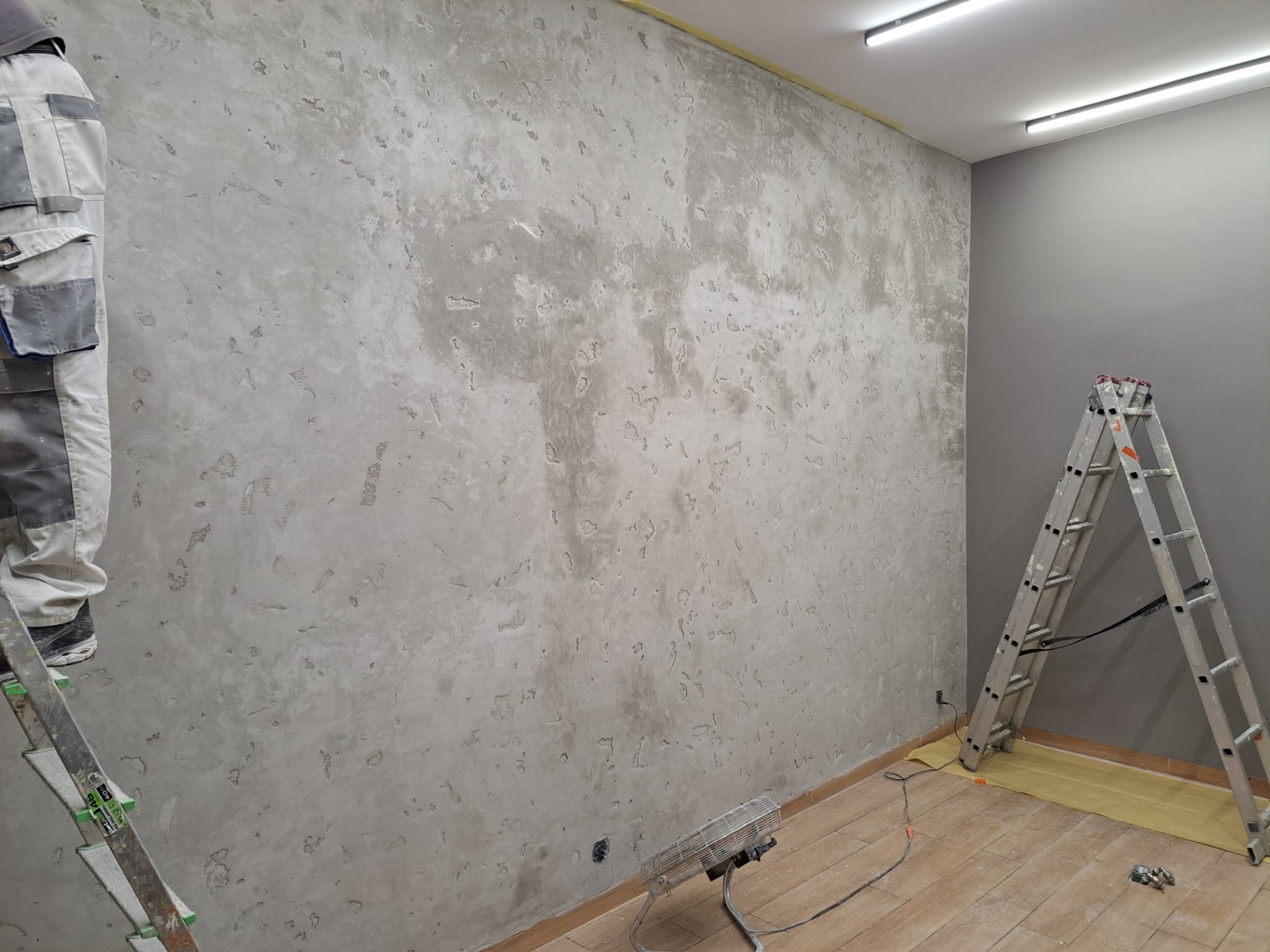 Ściana po nałożeniu dekoracyjnej, szarej struktury, widoczna drabina i fragment osoby w kombinezonie, podłoga zabezpieczona folią malarską.