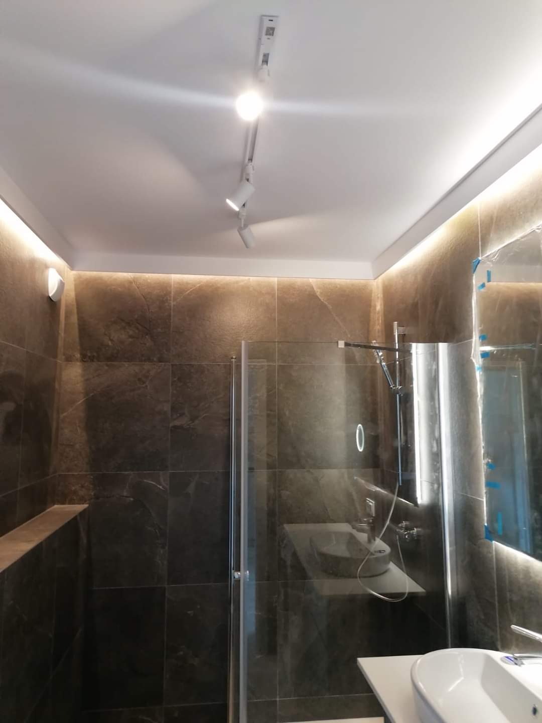 Nowoczesna łazienka z ciemnymi płytkami imitującymi marmur, przeszklonym prysznicem i oświetleniem punktowym na suficie.