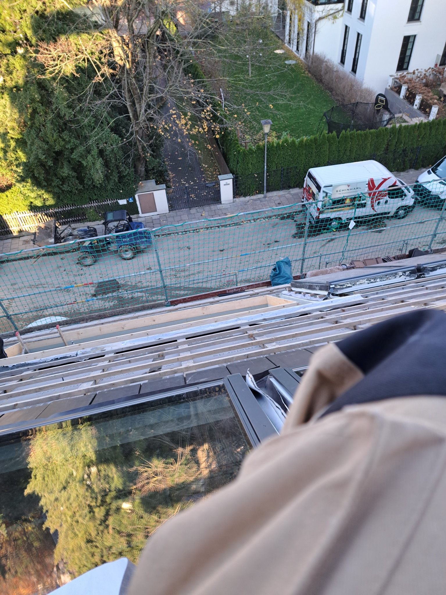 Widok z góry na konstrukcję dachu z drewnianymi belkami, zabezpieczoną siatką, z widocznym fragmentem pojazdu budowlanego i samochodu dostawczego firmy Schinharl w tle, z odbiciem drzew w szklanej...