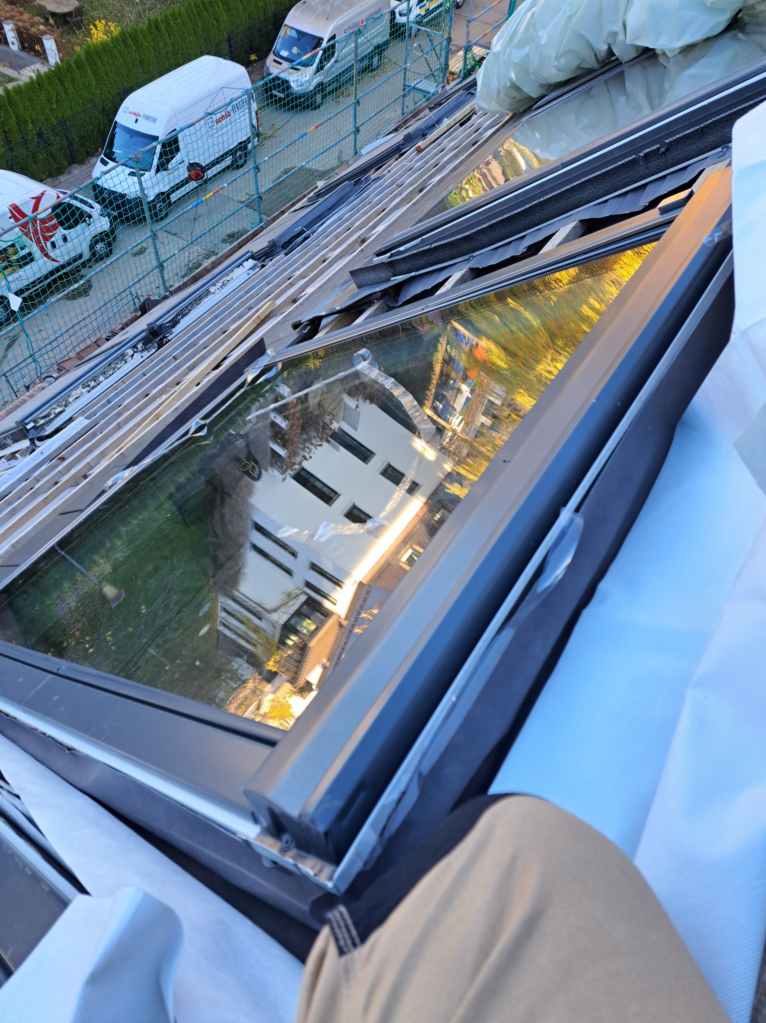 Montaż okna dachowego z widoczną konstrukcją dachu i odbiciem budynku w szybie, w tle zaparkowane samochody dostawcze firmy.