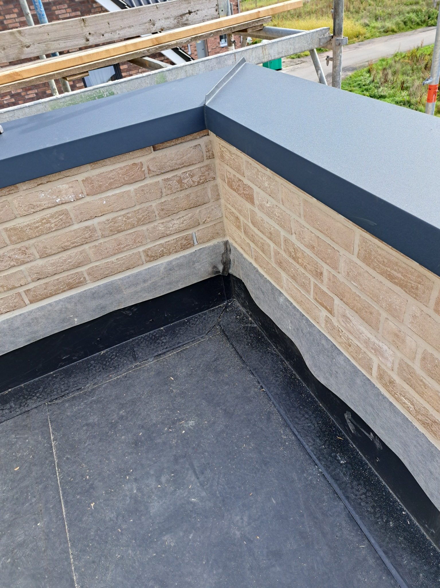 Widok z góry na fragment dachu z ceglaną ścianką attykową, zabezpieczoną szarą blachą, oraz czarną, bitumiczną izolacją przeciwwodną, z widocznym rusztowaniem w tle.