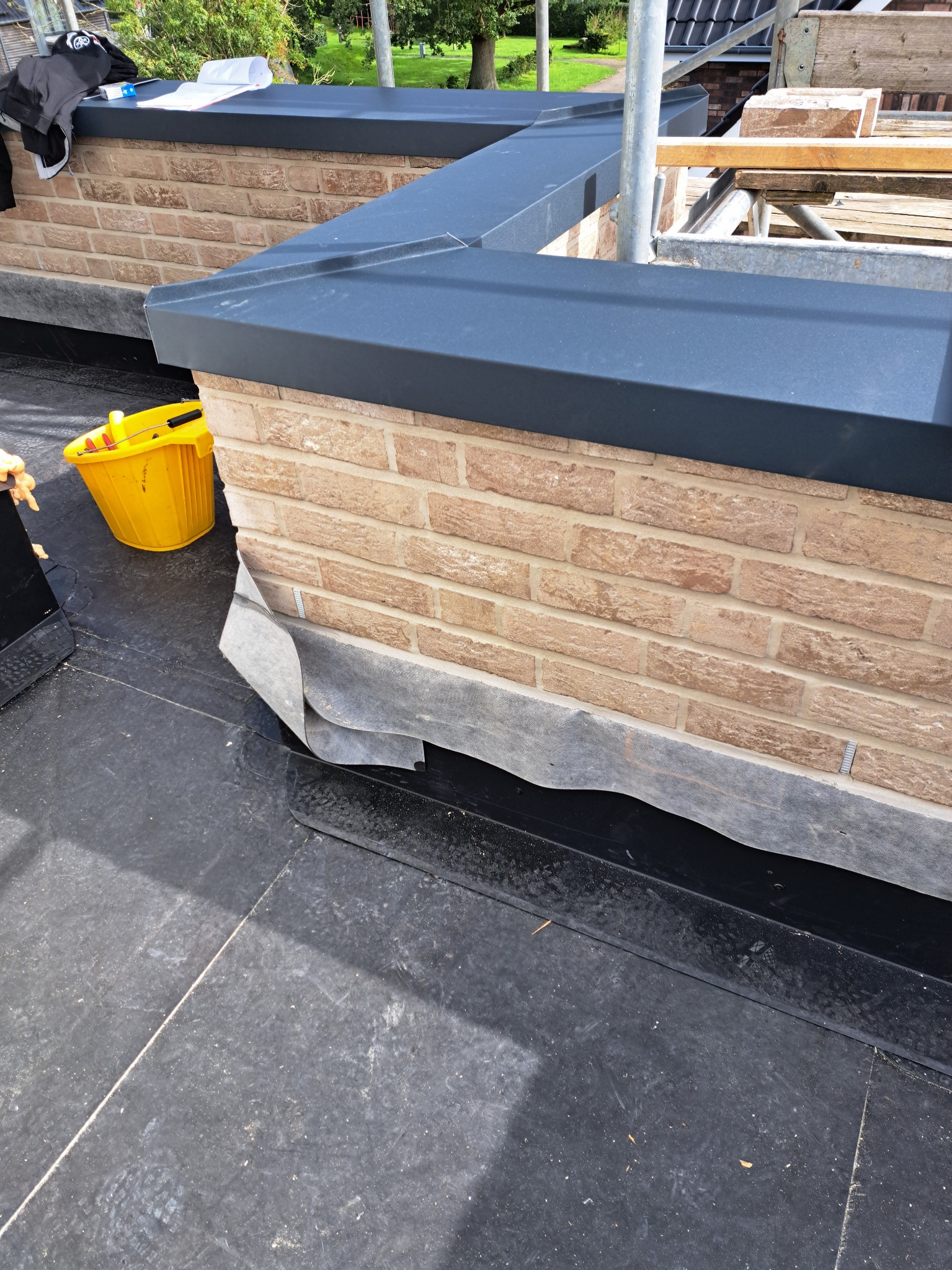 Murowana ściana z cegły klinkierowej z czarnym obramowaniem, widoczna żółta wiaderko z narzędziami i fragment rusztowania. Na dole folia zabezpieczająca. Praca w toku.