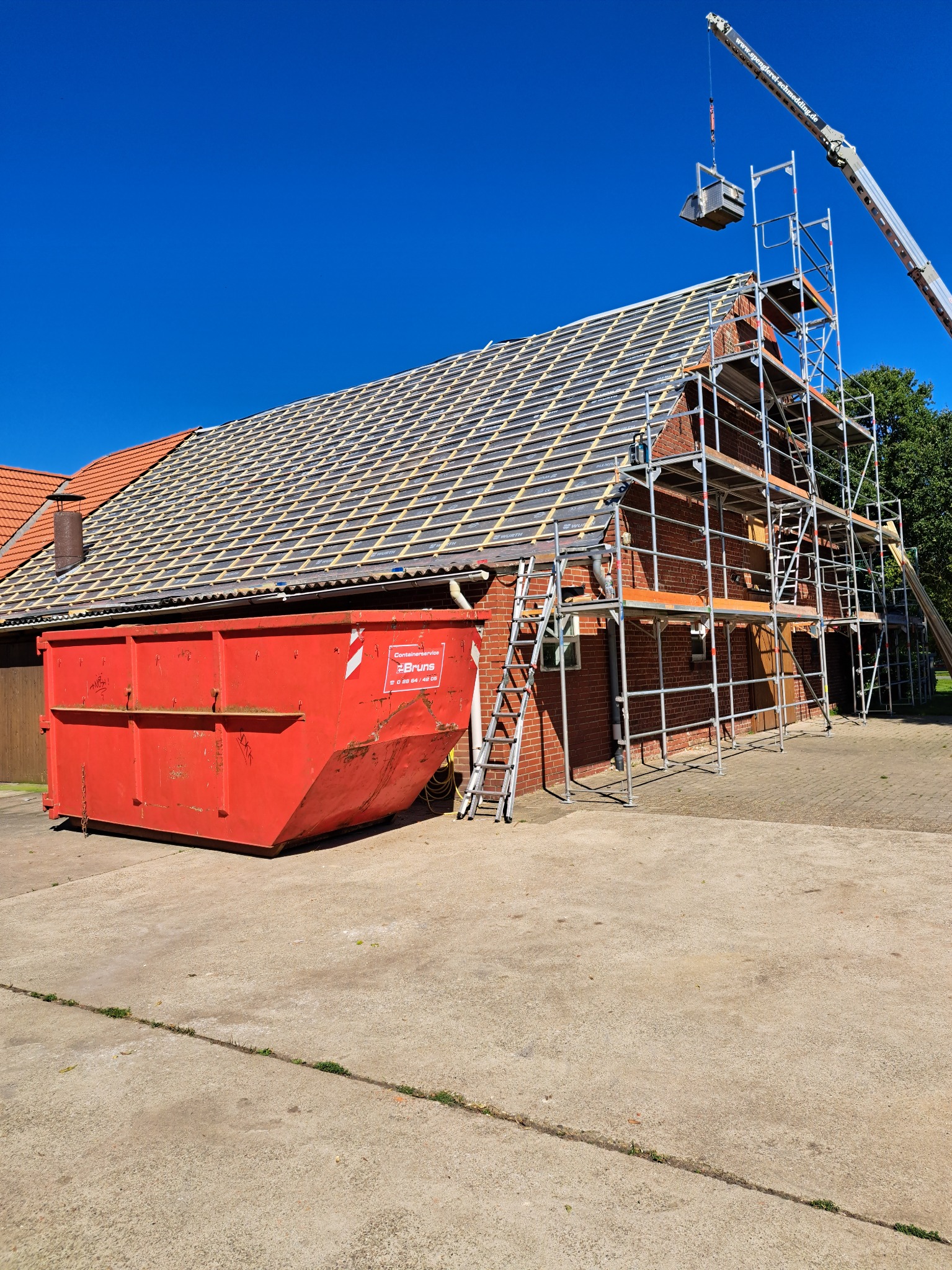 Czerwony kontener na gruz obok budynku z cegły z rusztowaniem i dźwigiem podczas wymiany dachu z dachówki ceramicznej.