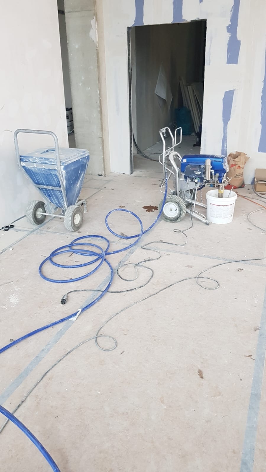 Przygotowane do malowania wnętrze z agregatem malarskim i wózkiem na farbę, widoczne przewody i worki z materiałami budowlanymi.