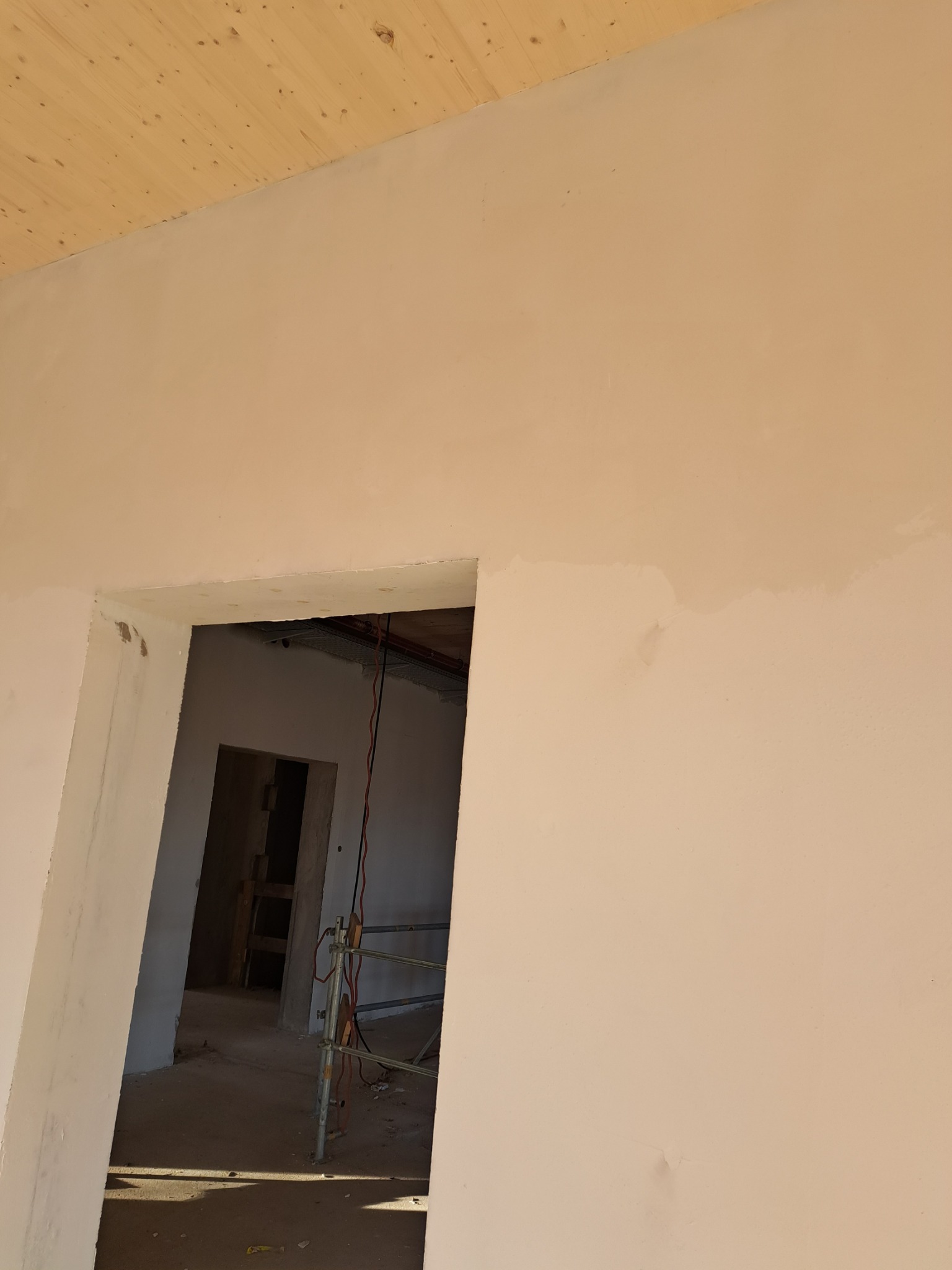 Świeżo otynkowana ściana w odcieniach beżu z widocznym fragmentem drewnianego sufitu i prześwitem do kolejnego pomieszczenia, gdzie widoczne są elementy rusztowania.