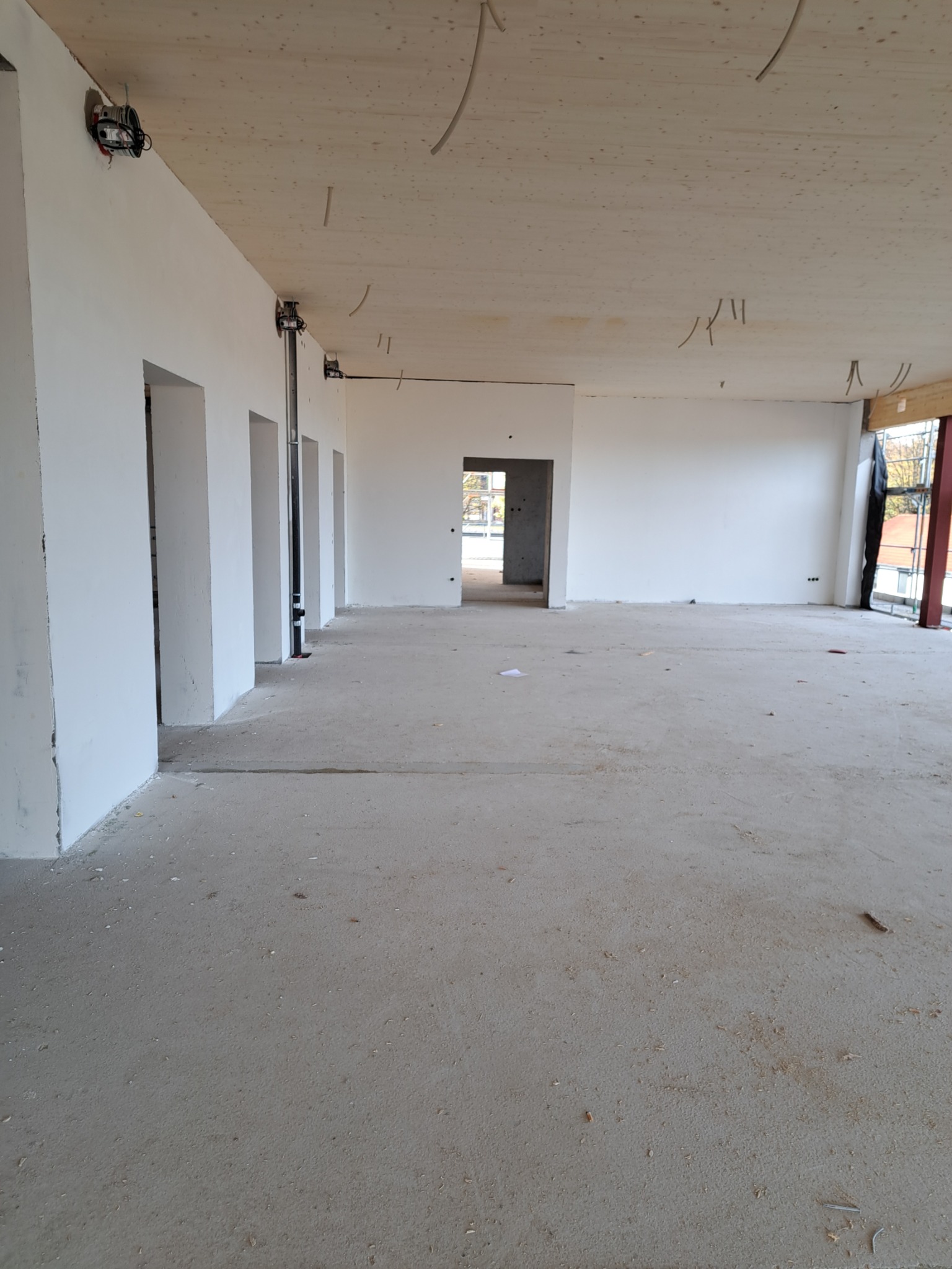 Wnętrze w stanie deweloperskim z surową podłogą, białymi ścianami i drewnianym stropem, widoczne wystające kable i instalacje.