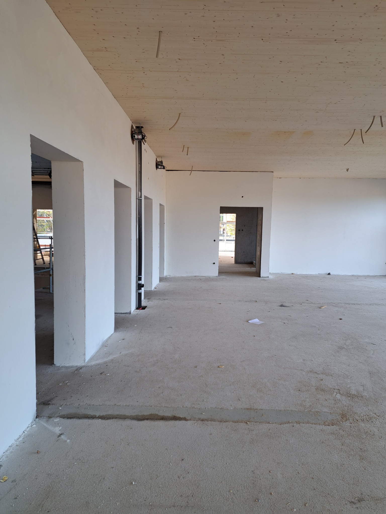 Wnętrze w trakcie prac wykończeniowych: surowy beton na podłodze, świeżo pomalowane białe ściany z widocznymi otworami drzwiowymi i drewniany sufit z instalacją elektryczną.