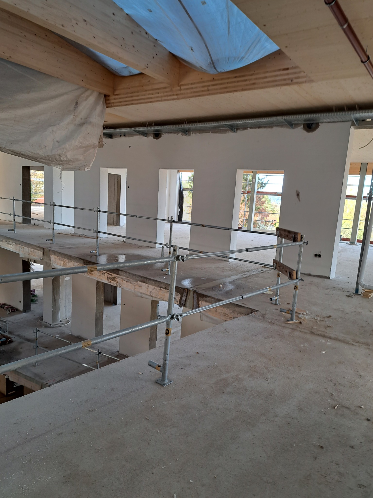 Wnętrze w trakcie prac budowlanych: surowy beton, drewniane belki stropowe pod przykryciem z folii, metalowe barierki zabezpieczające, widoczne okna i otwory drzwiowe w surowych ścianach.